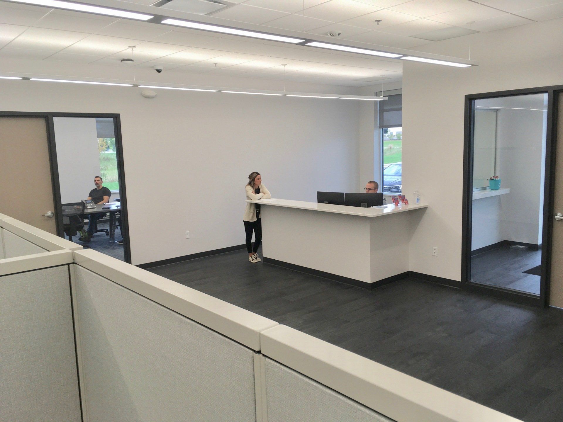 Reception area with three people: two at the desk, one in a nearby office. White desk, dark floor.