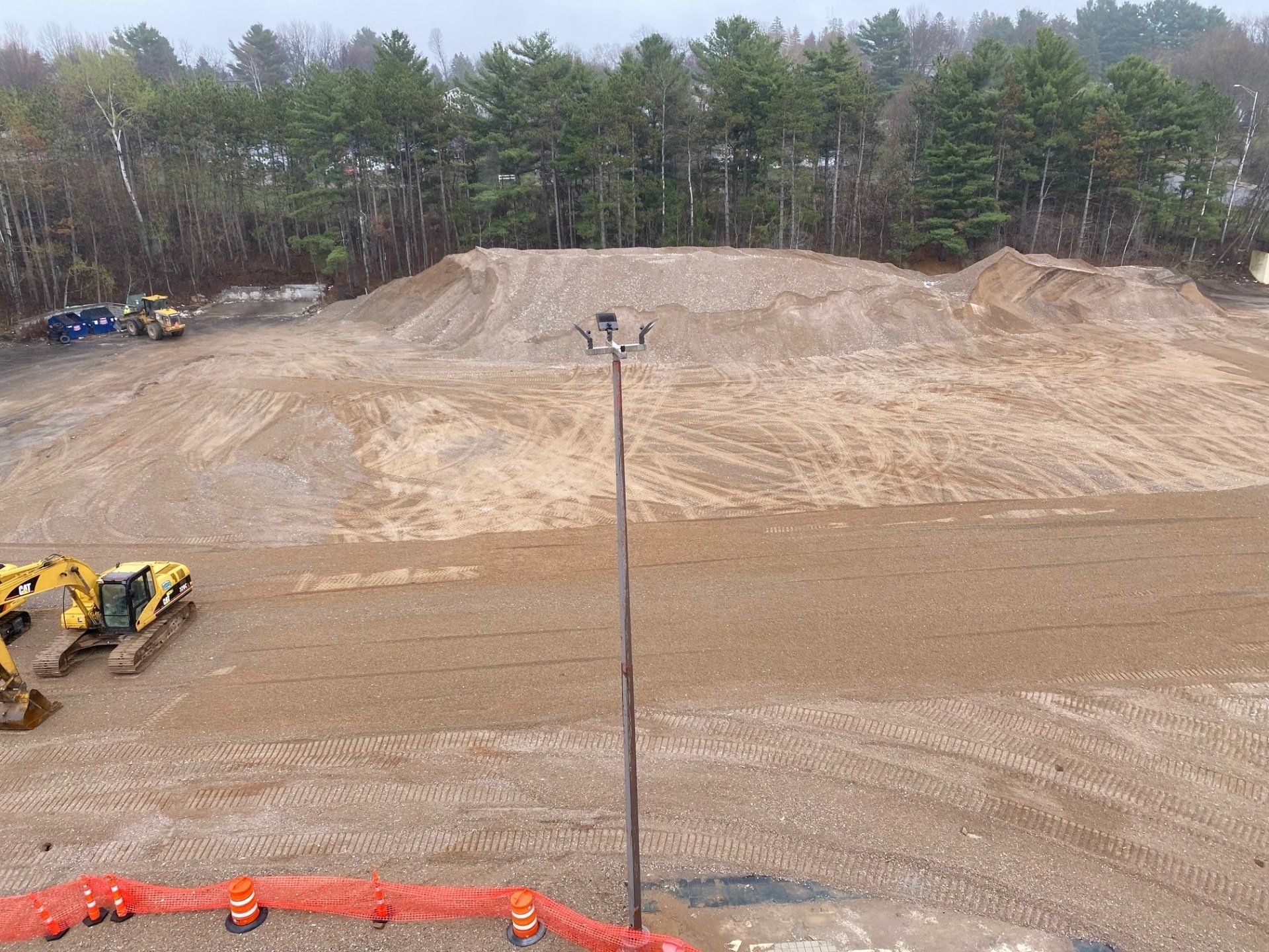 Construction site with large dirt piles, heavy machinery, and trees in the background.