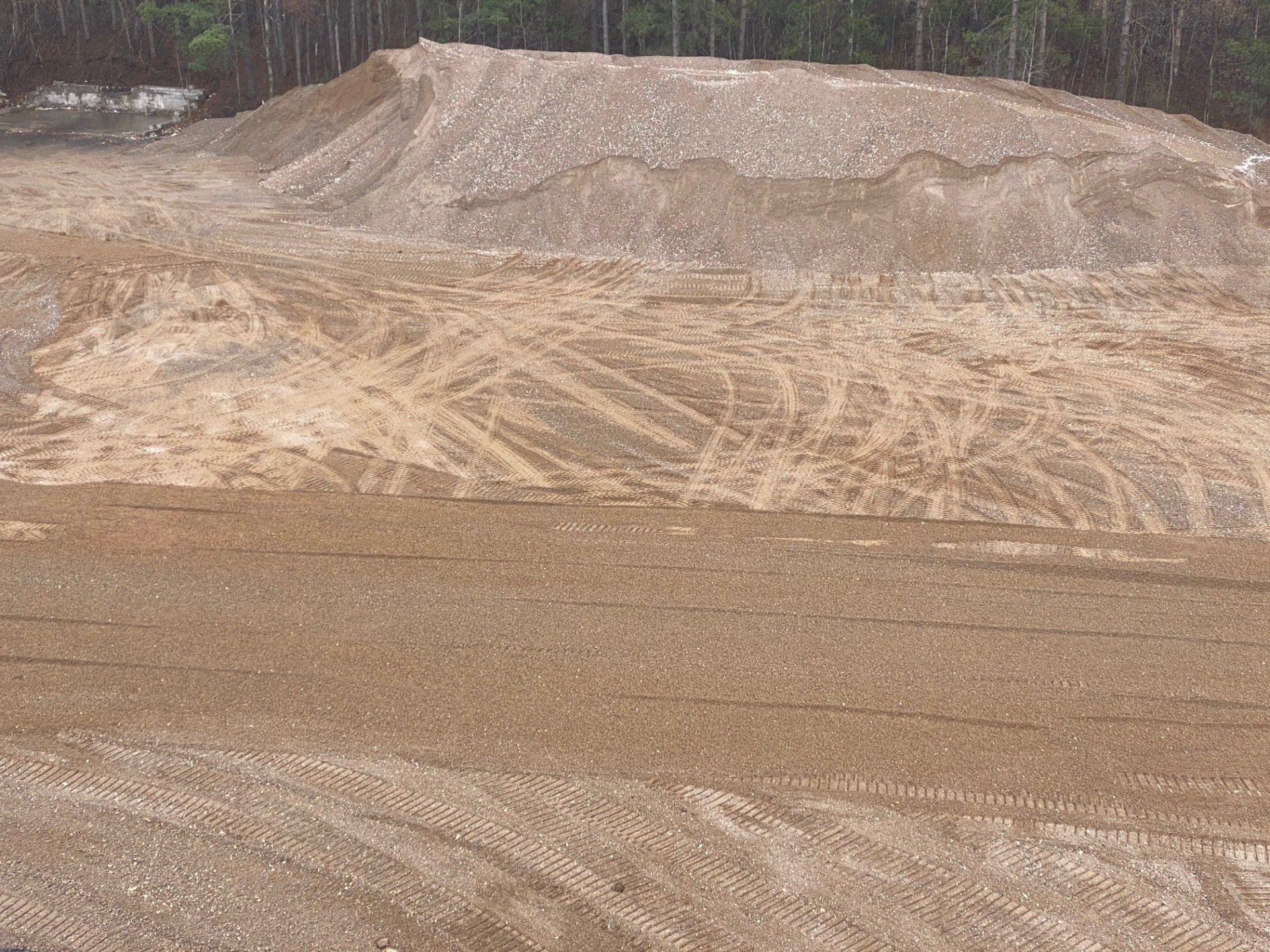 A large pile of gravel with tire tracks on a graded, gravel surface. Wooded area in the background.