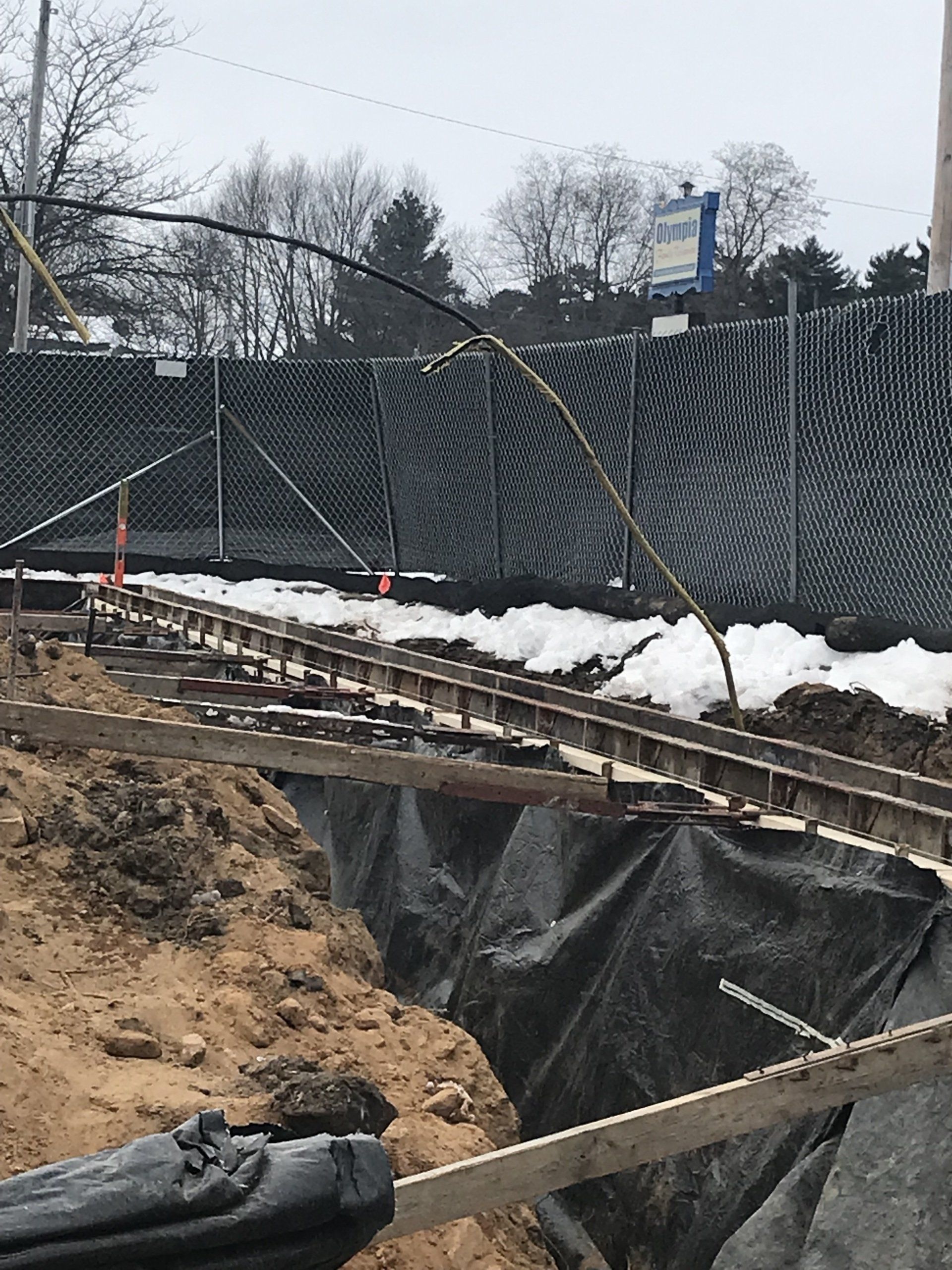 Construction site with a trench, wooden supports, black liner, and a fence. Utility lines overhead.