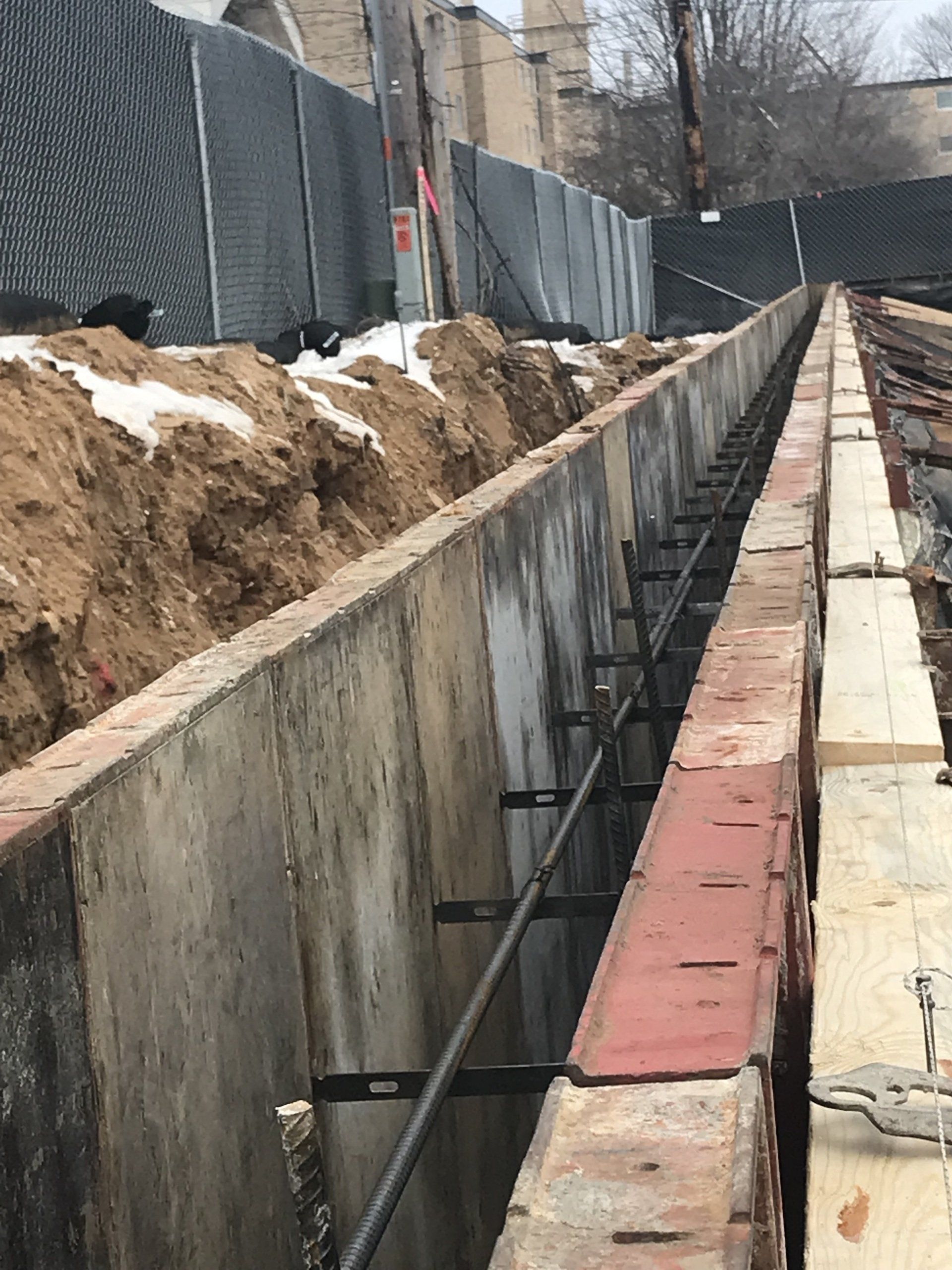 Construction site with a long trench, concrete walls, and rebar reinforcement. Dirt and snow present.
