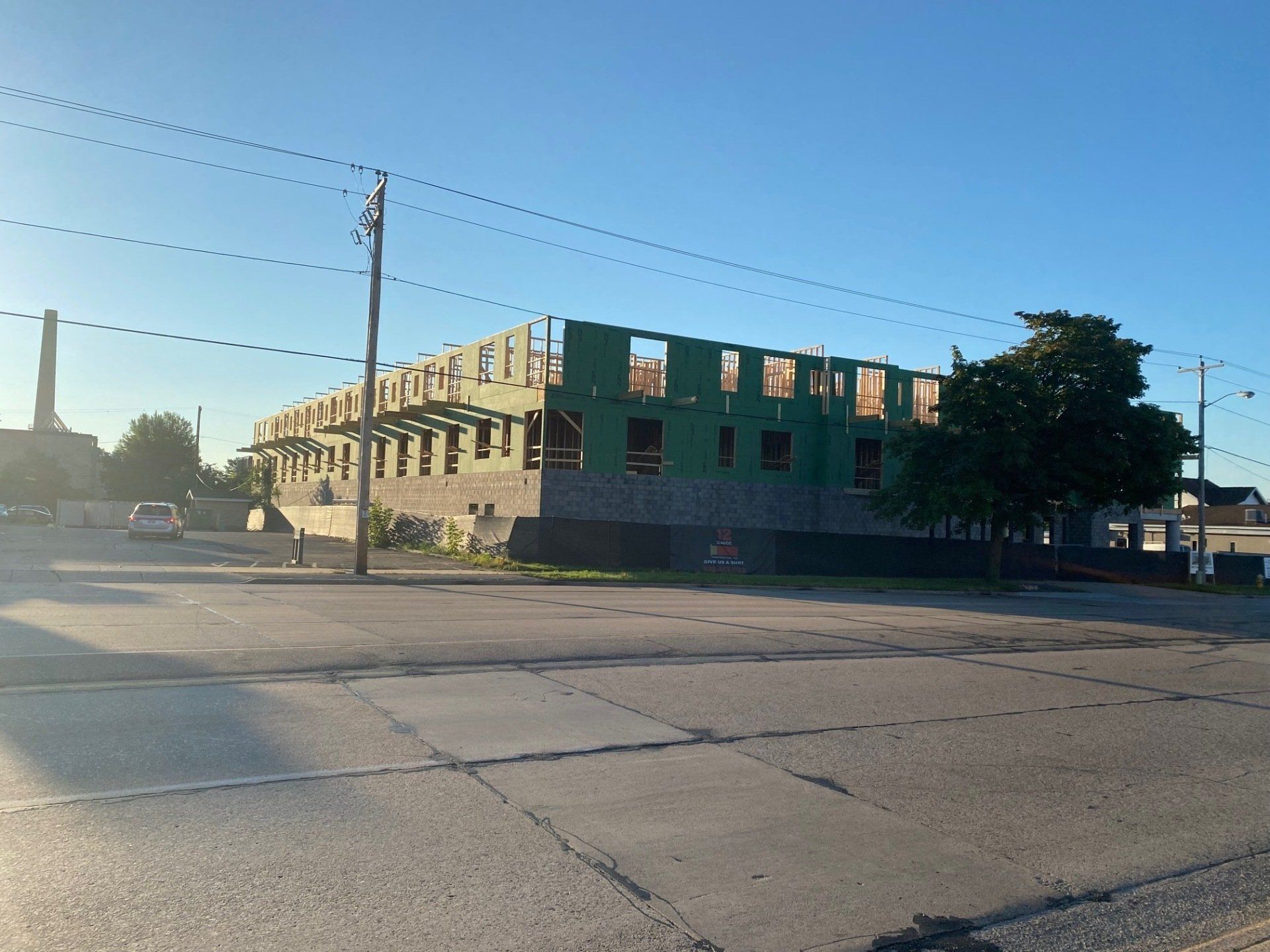 Construction of a multi-story building frame on a street corner, with power lines overhead and a factory chimney in the distance.