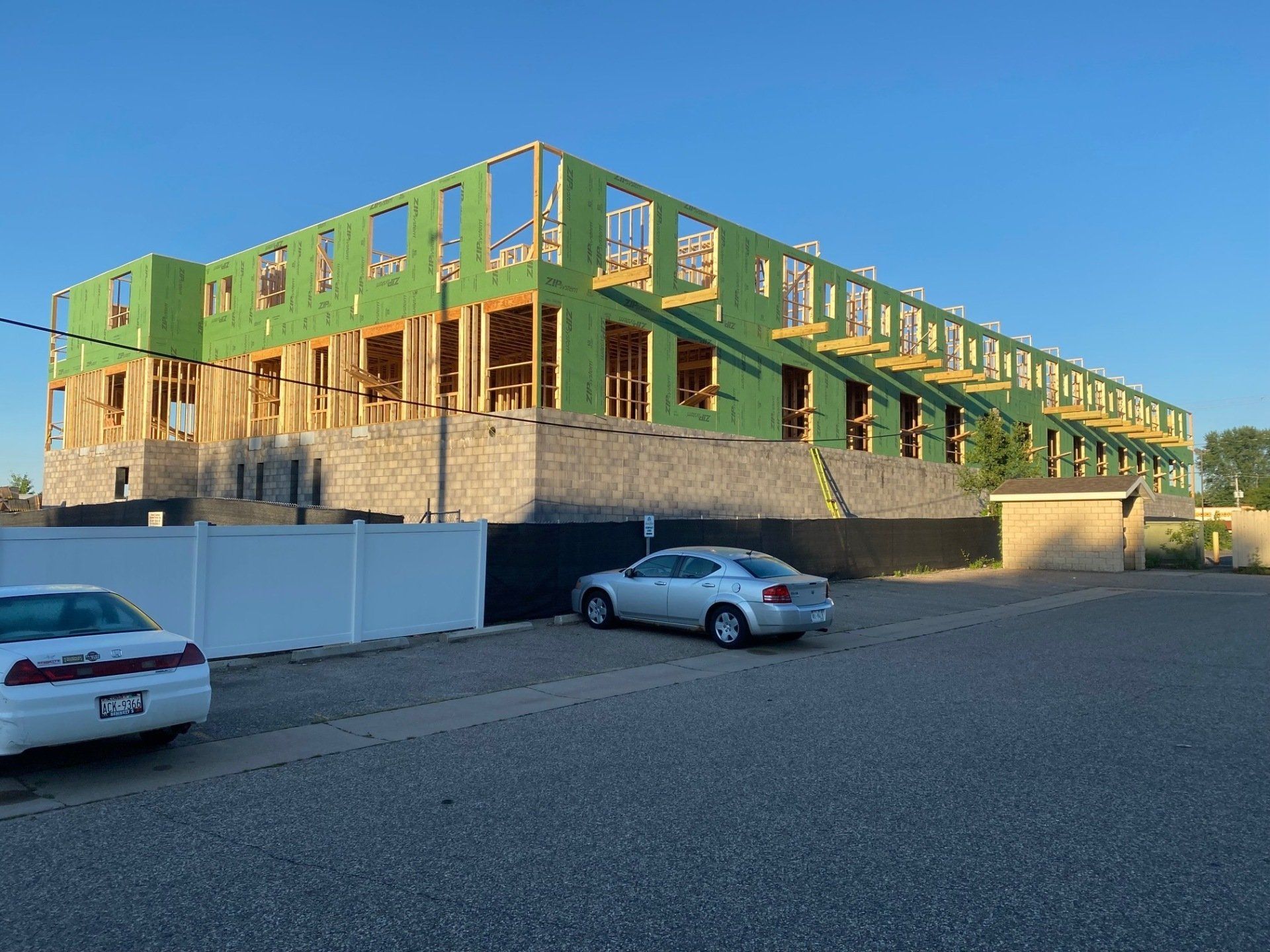 Building under construction; wood framing and green sheathing. Cars parked in front on a sunny day.
