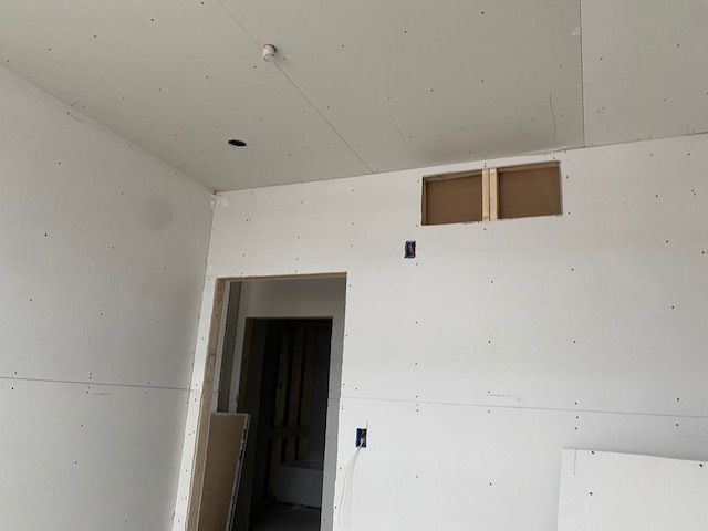 Interior view of a room under construction with drywall installed. Features a doorway, vents, electrical outlets, and a smoke detector.