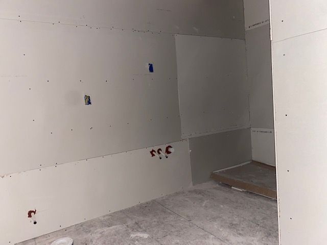 Interior room under construction, with drywall walls and a concrete floor. Electrical and plumbing fixtures visible.
