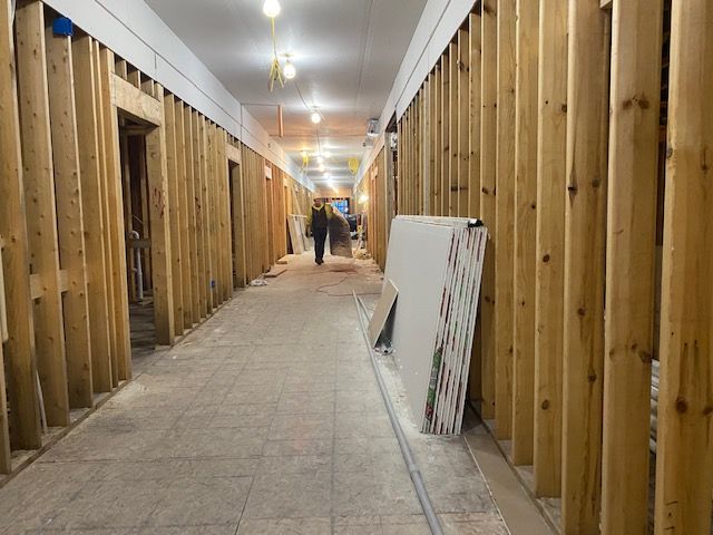 Hallway under construction with exposed wooden studs. A person walks in the distance. Drywall sheets lean against the wall.