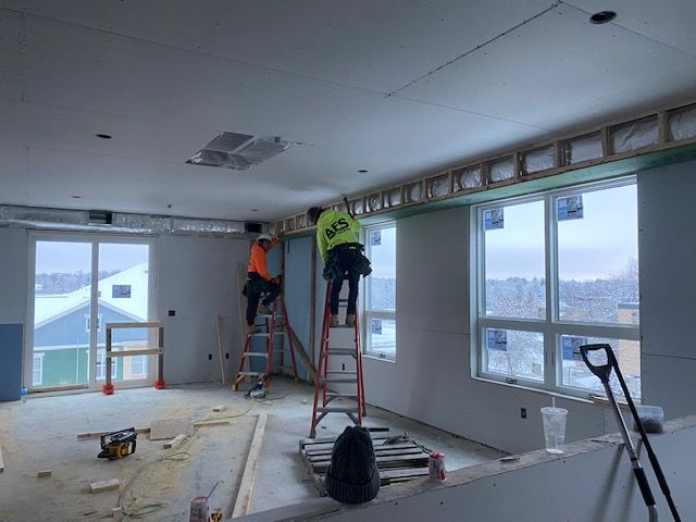 Construction workers installing drywall in a room with windows overlooking a winter landscape.