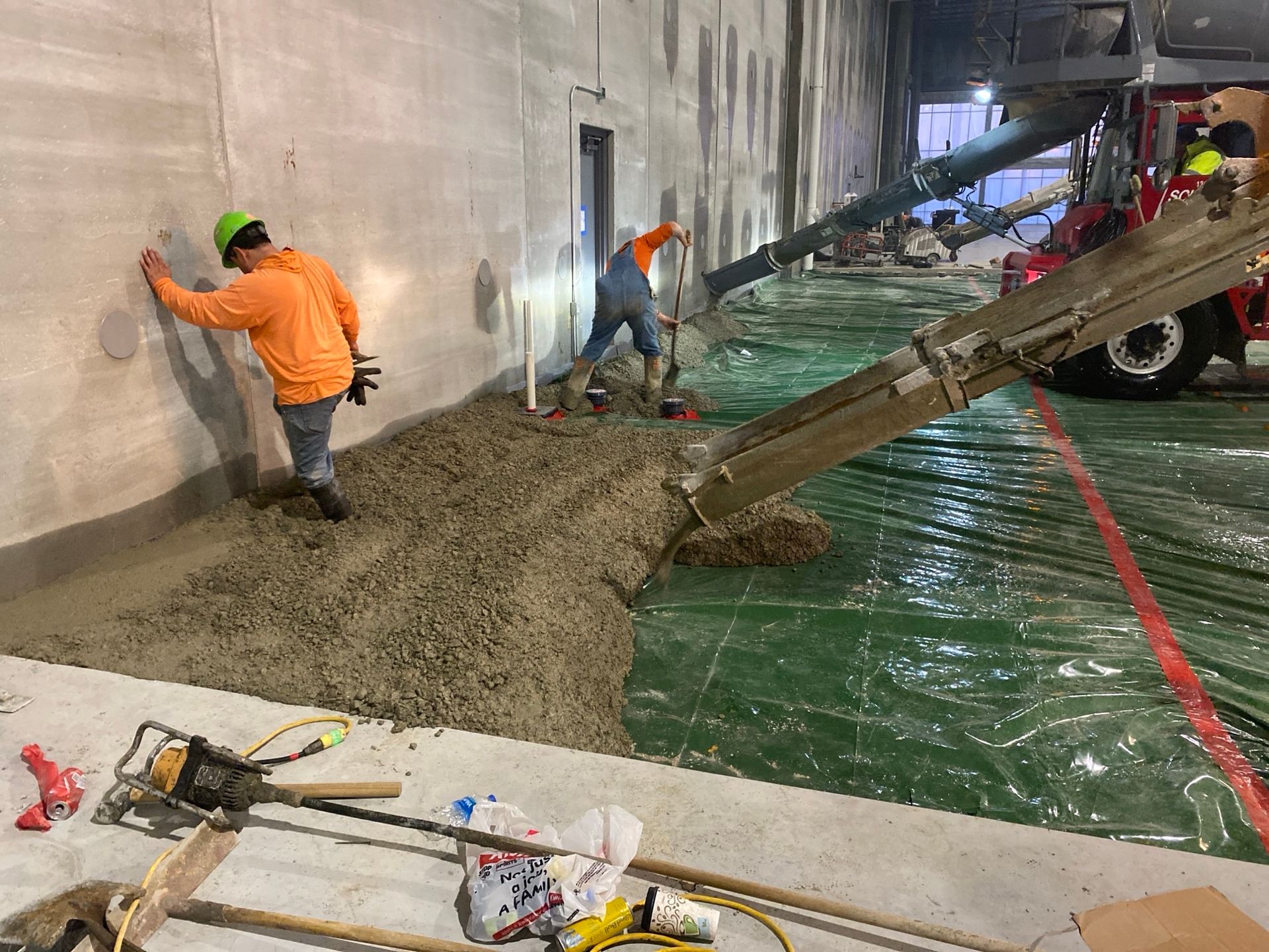 Construction workers pouring concrete on a green tarp. One worker checks the wall, another guides the flow.