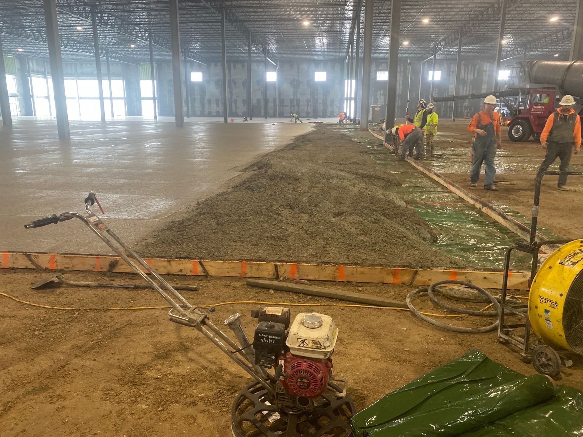 Construction workers pouring concrete on a large indoor floor.  A walk-behind machine, hose, and trucks are present.