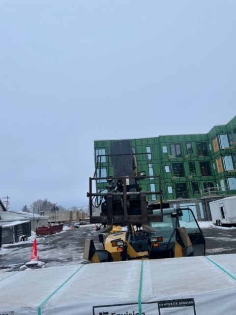 Forklift on construction site in front of a partially-built green building on a snowy day.