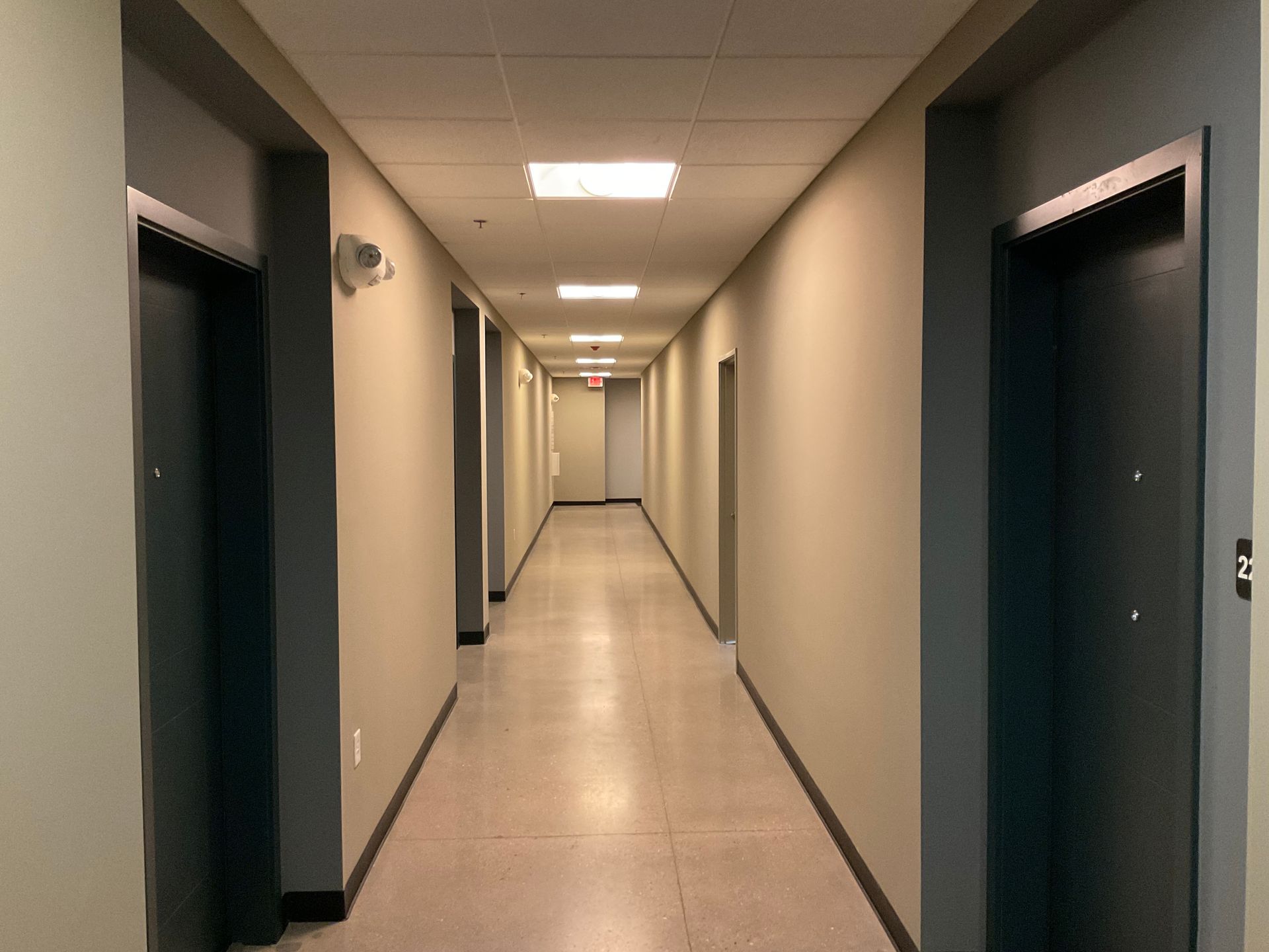 Long hallway with beige walls, gray doors, and bright overhead lights.
