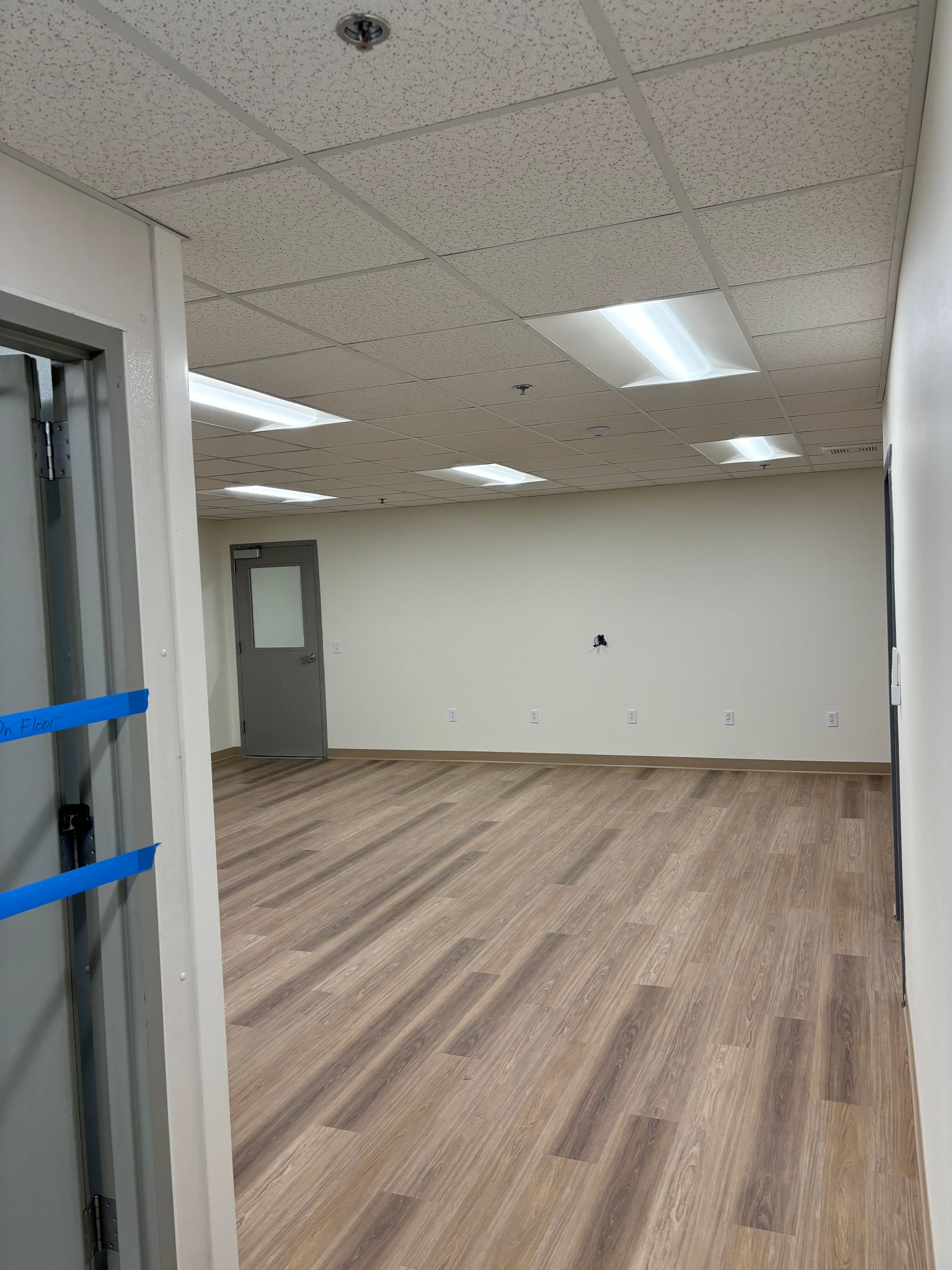 Empty room with wood-look flooring, white walls, and a drop ceiling with fluorescent lights. A gray door is in the back.