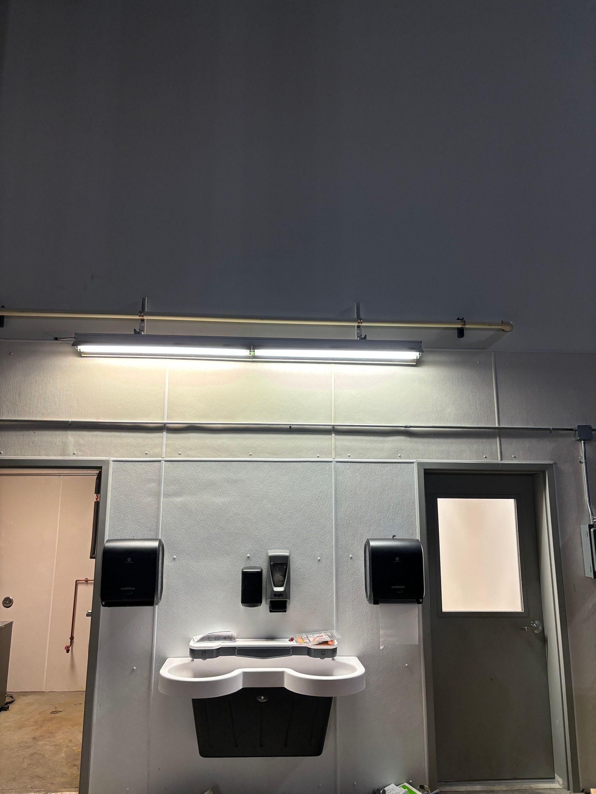 Bathroom with a sink, soap and paper towel dispensers, and two doors. White and gray walls and a fluorescent light.