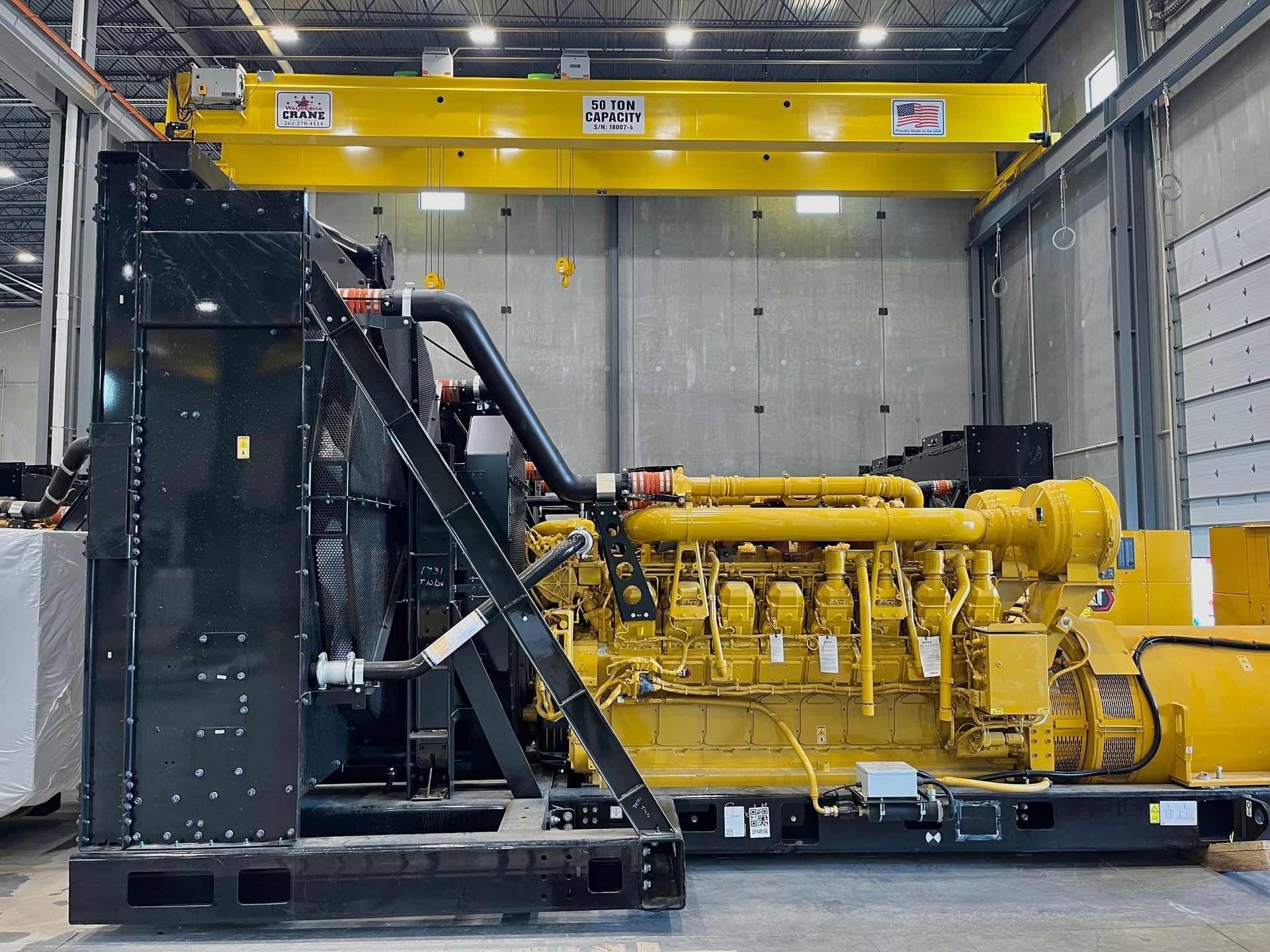 Yellow industrial generator in a factory setting, with a black frame and overhead crane.