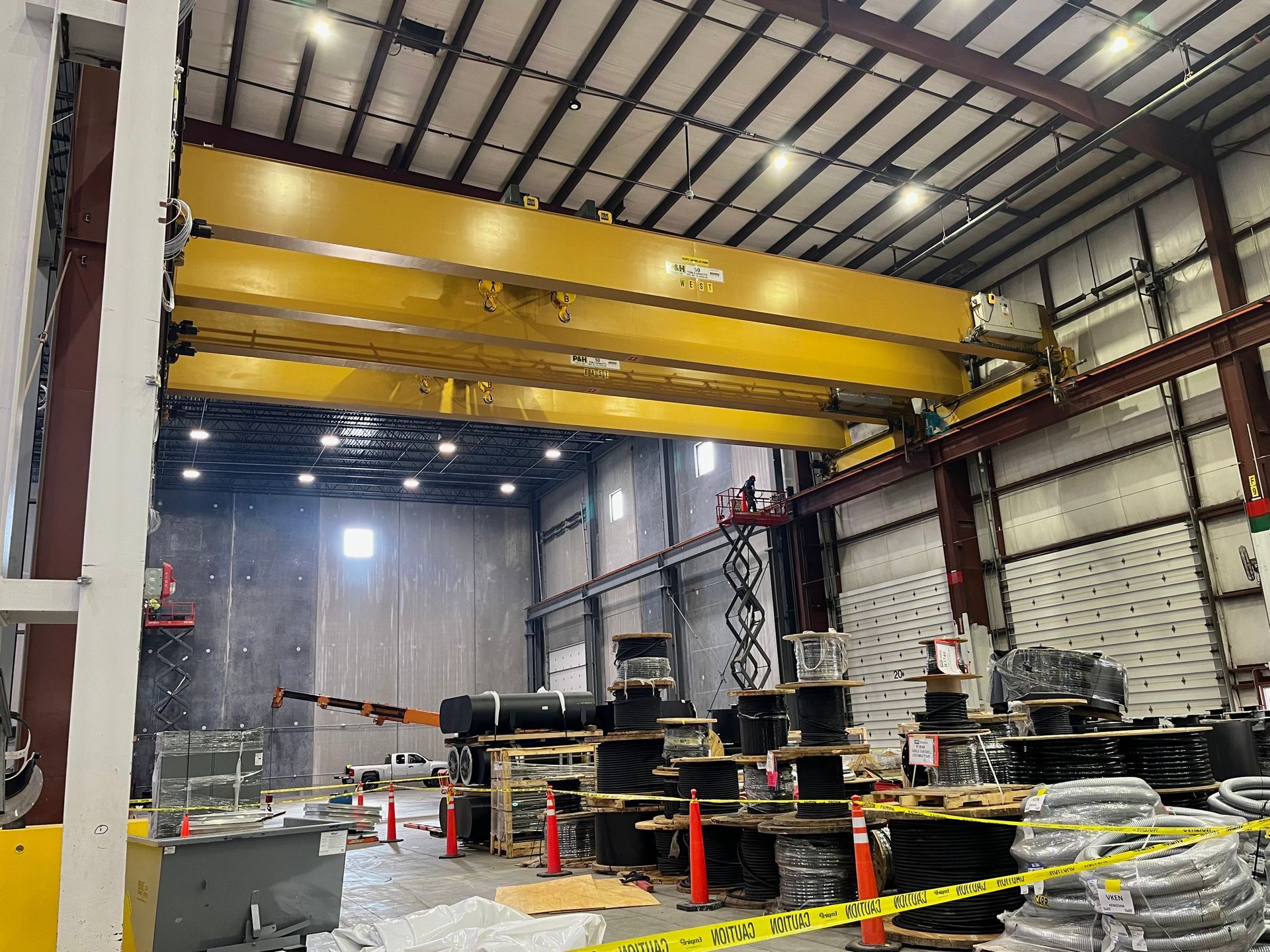 Yellow overhead crane in a large warehouse. Cables and spools sit on pallets on the floor.