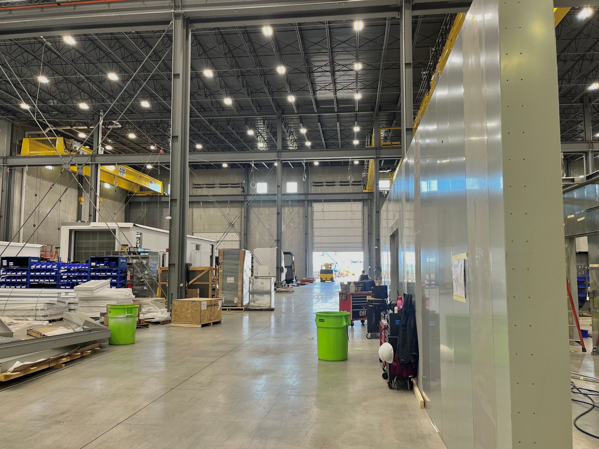 Large industrial warehouse interior with open bay door. Green bins and equipment line the floor, overhead crane in background.
