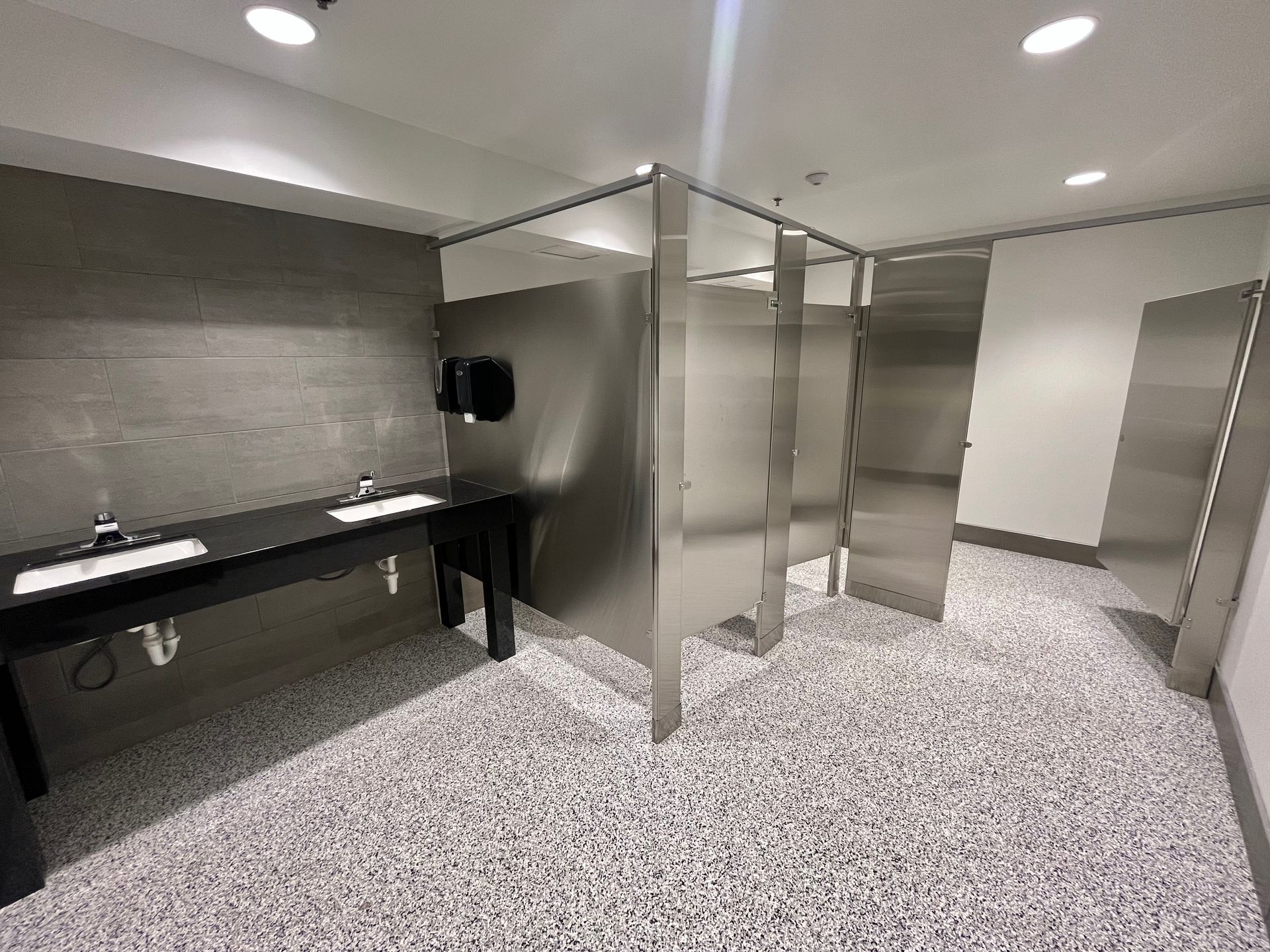 Modern public restroom with stainless steel stalls, sinks, and speckled flooring.