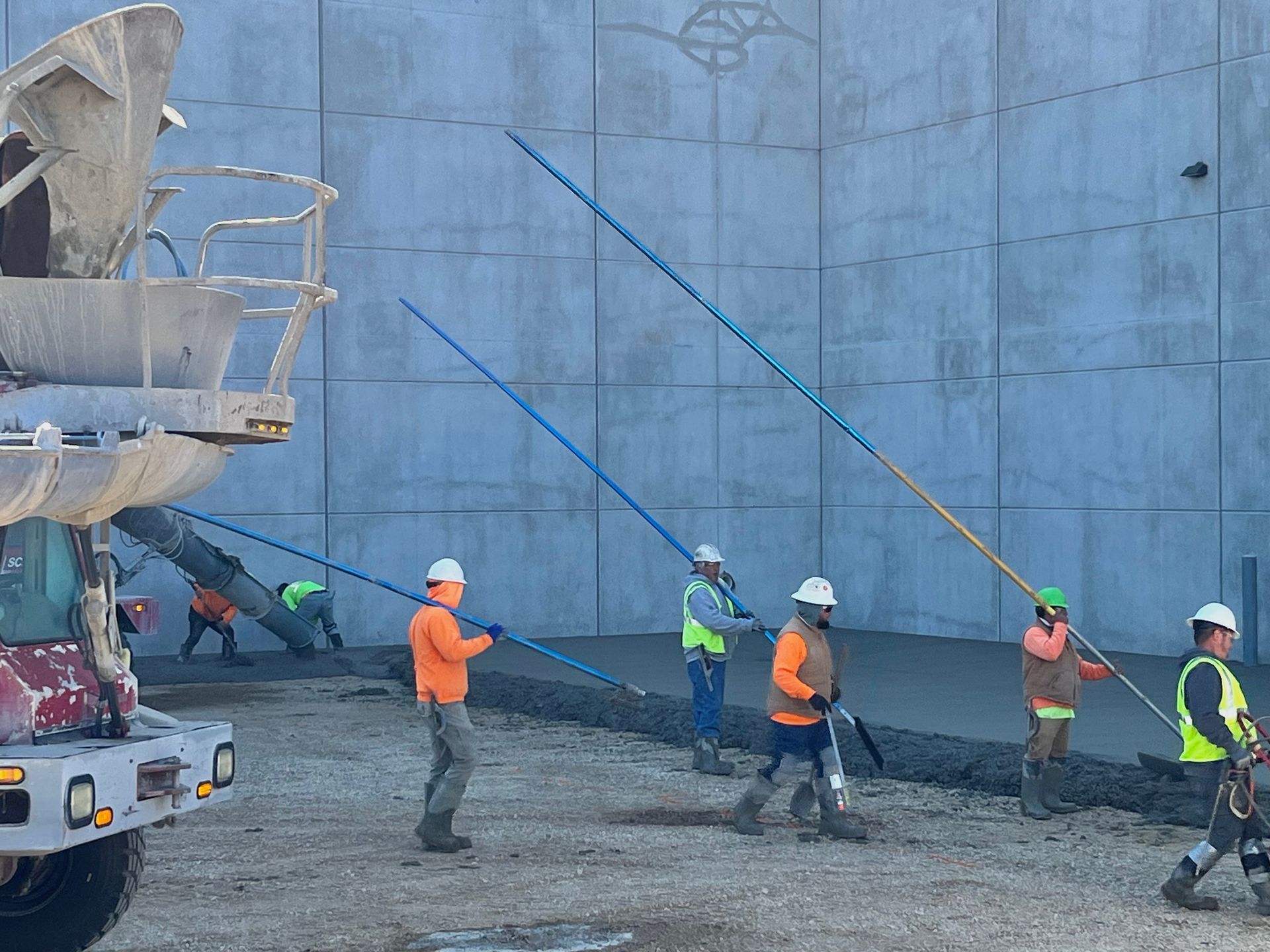 Construction workers pouring concrete, using long poles to level it. Gray walls, sunny outdoors.