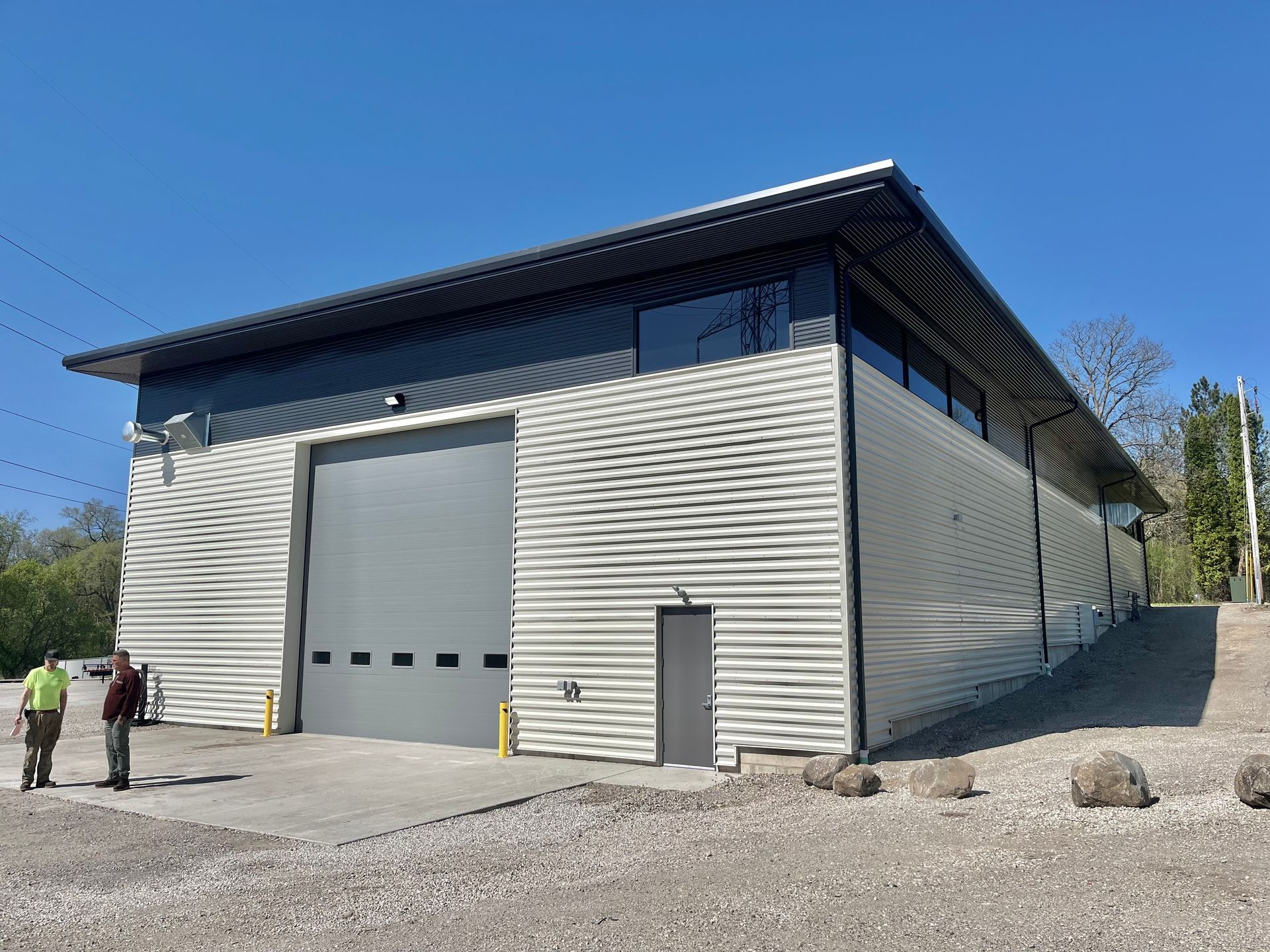 A gray industrial building with a large garage door. Two people stand near the entrance under a clear, blue sky.