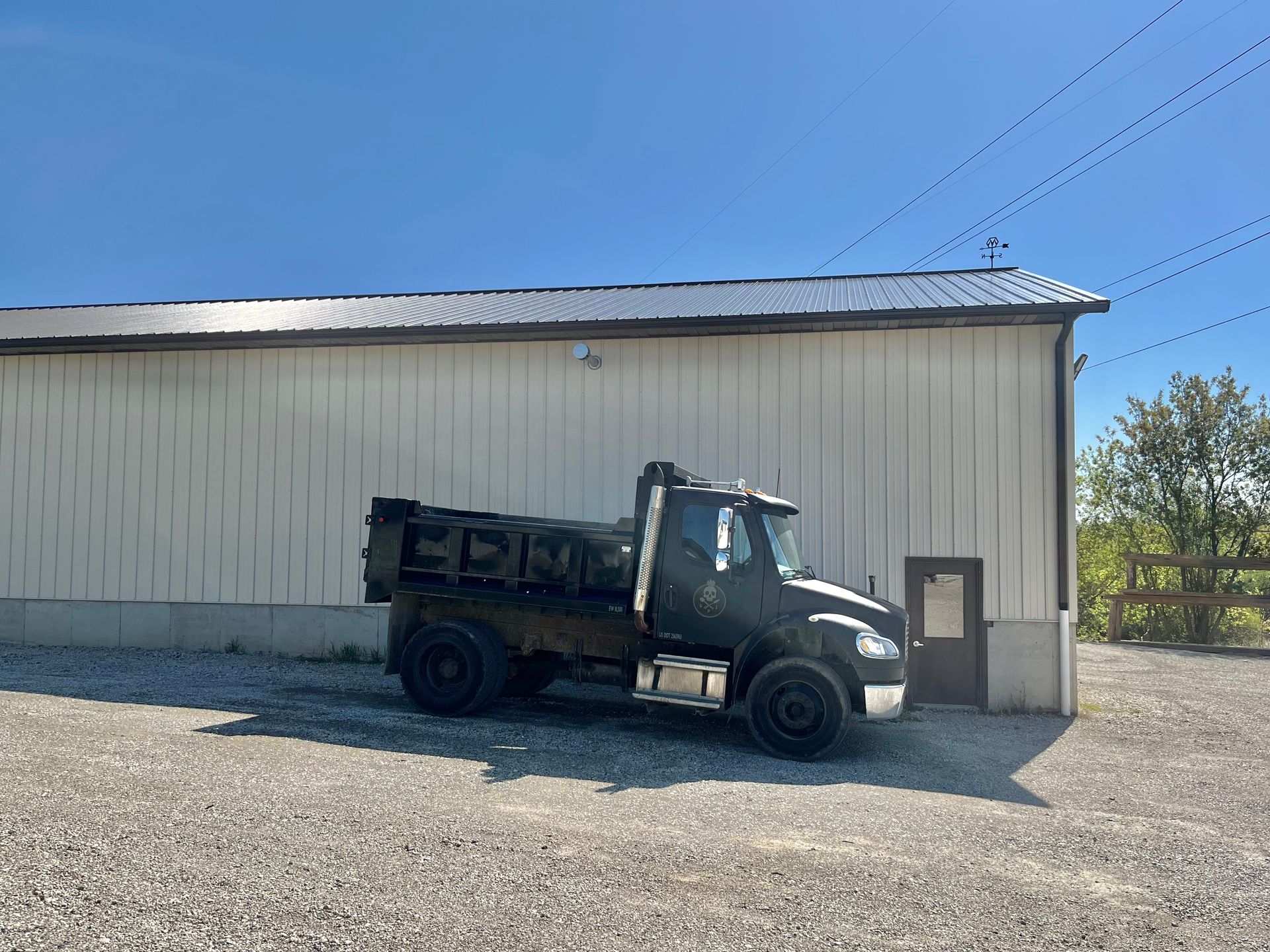 Dark dump truck parked in front of a white metal building on a gravel lot.