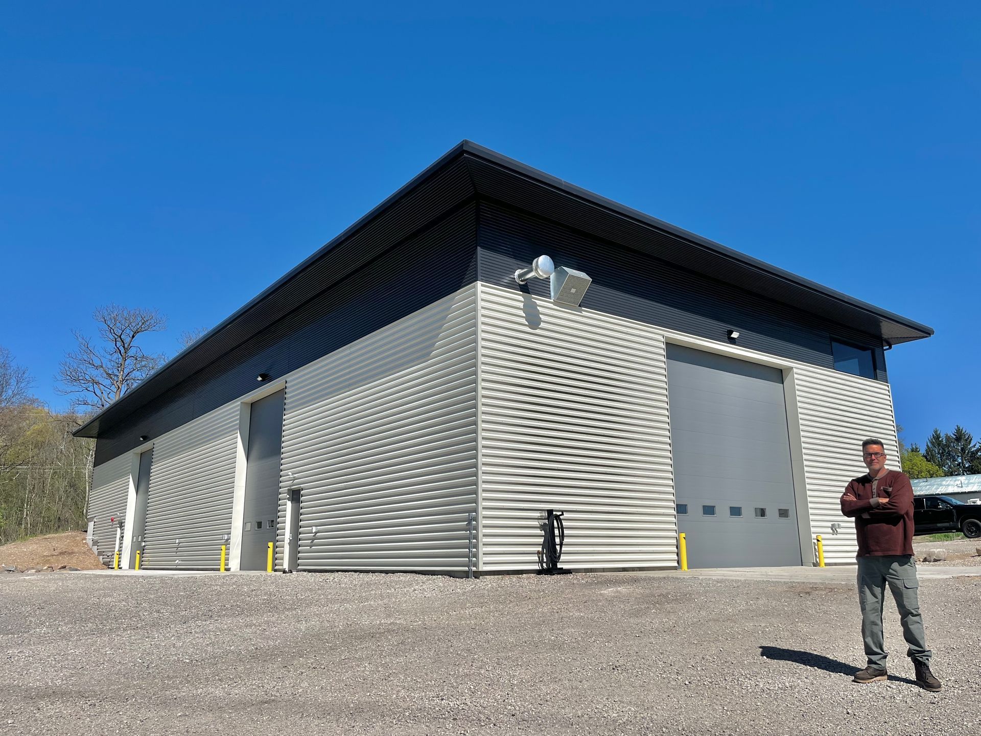 Man standing outside a corrugated metal building with large garage doors and solar panels, under a blue sky.