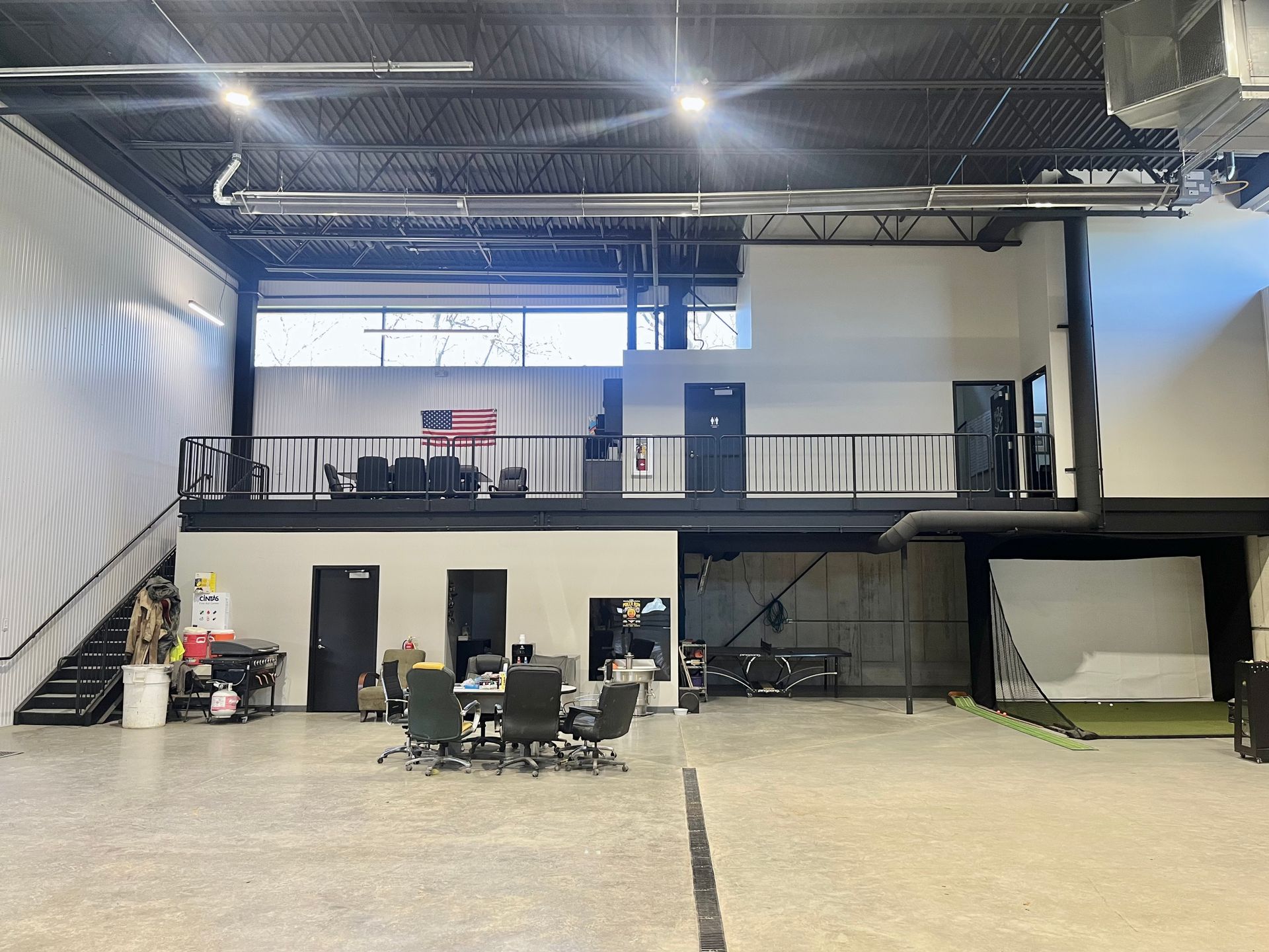 Interior of a modern space with a mezzanine, golf simulator, and seating area. Concrete floor, high ceilings.