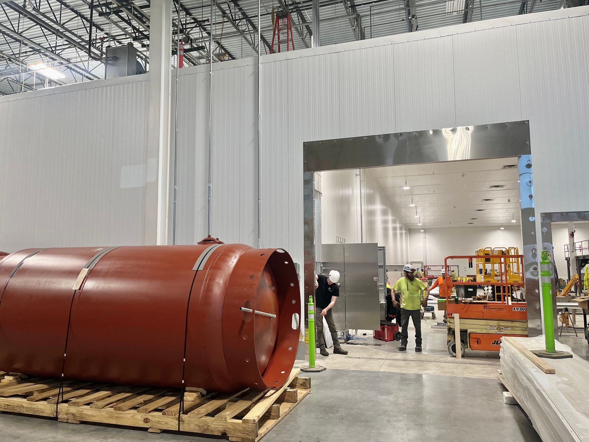 Large red cylindrical tank on a pallet inside a facility. Workers near a doorway into a bright interior.