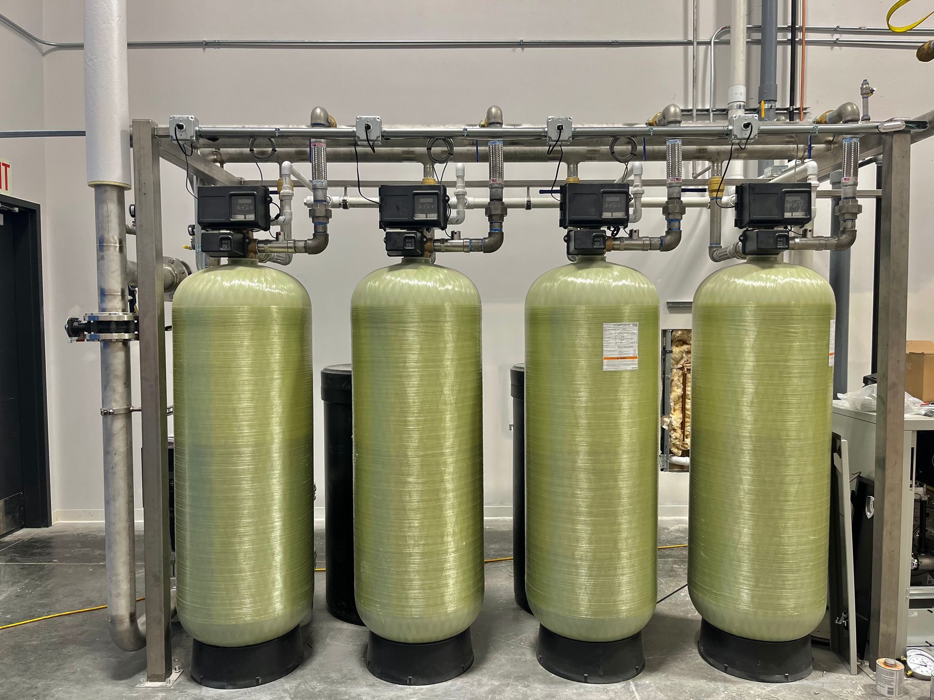 Four tall, cylindrical water filter tanks with control boxes, linked by pipes, in a room.