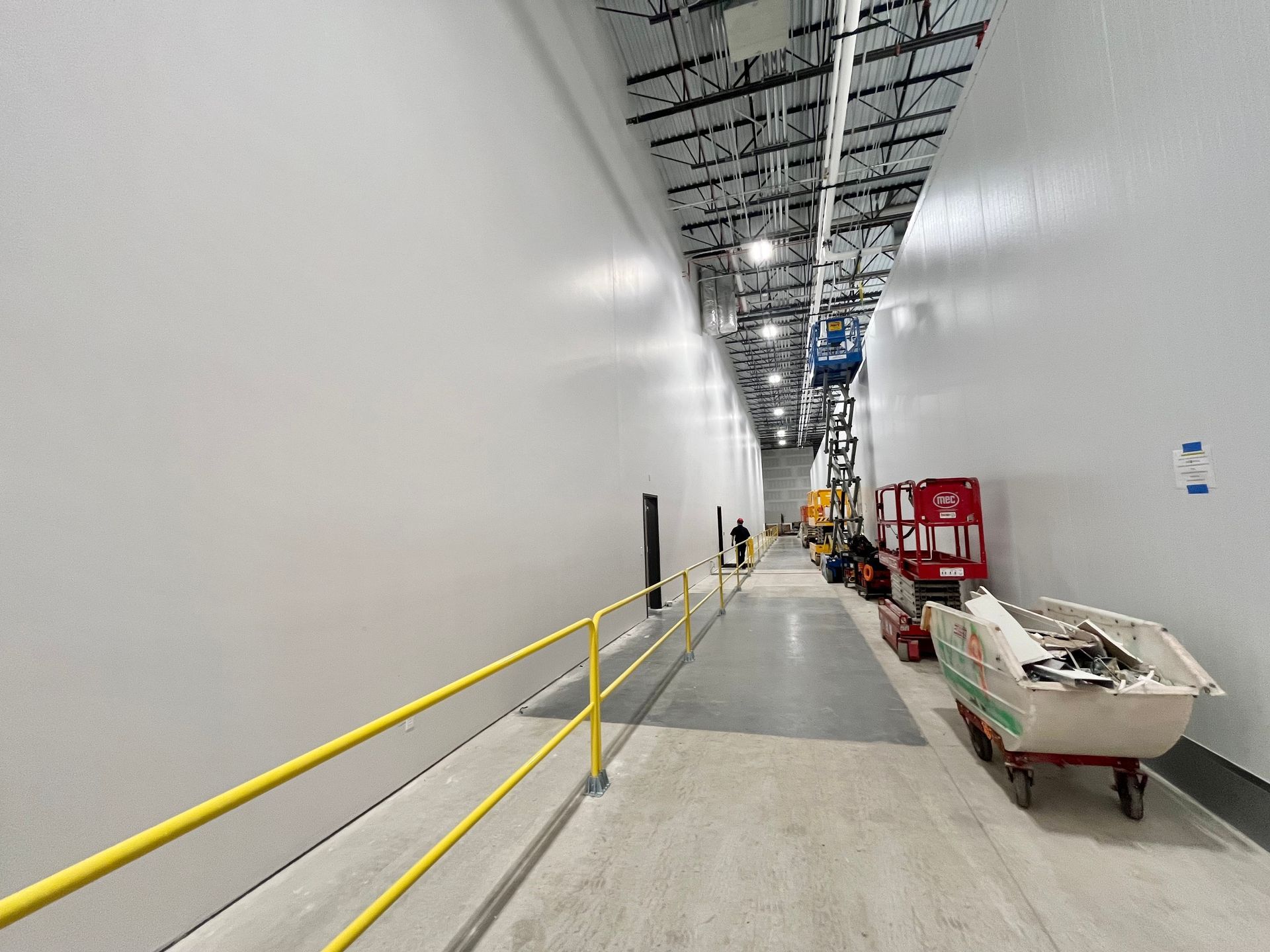 Long, narrow warehouse hallway with white walls, yellow railing, and a person walking in the distance.