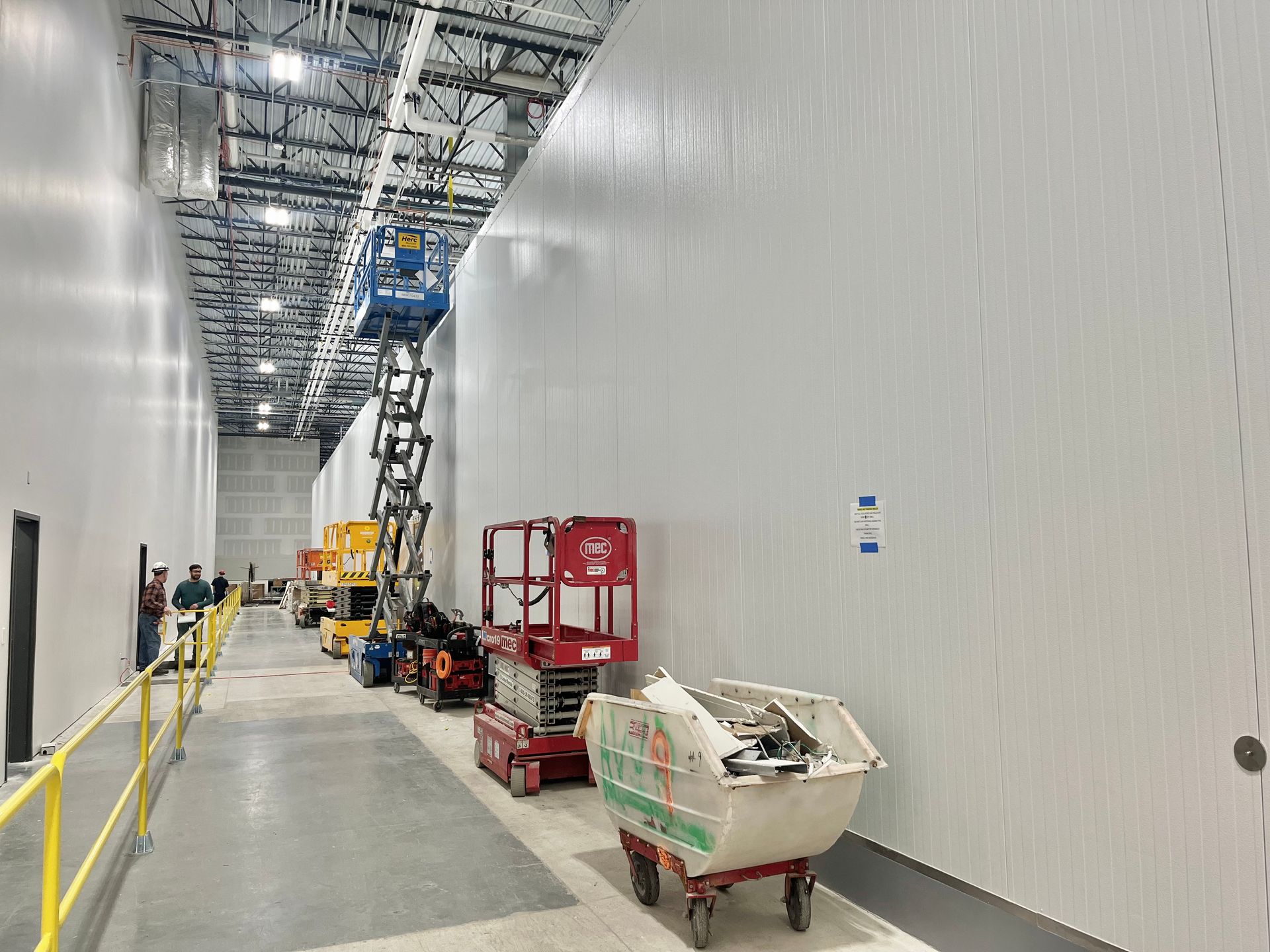 Construction site: Workers near tall white walls and equipment in a large, open industrial space.