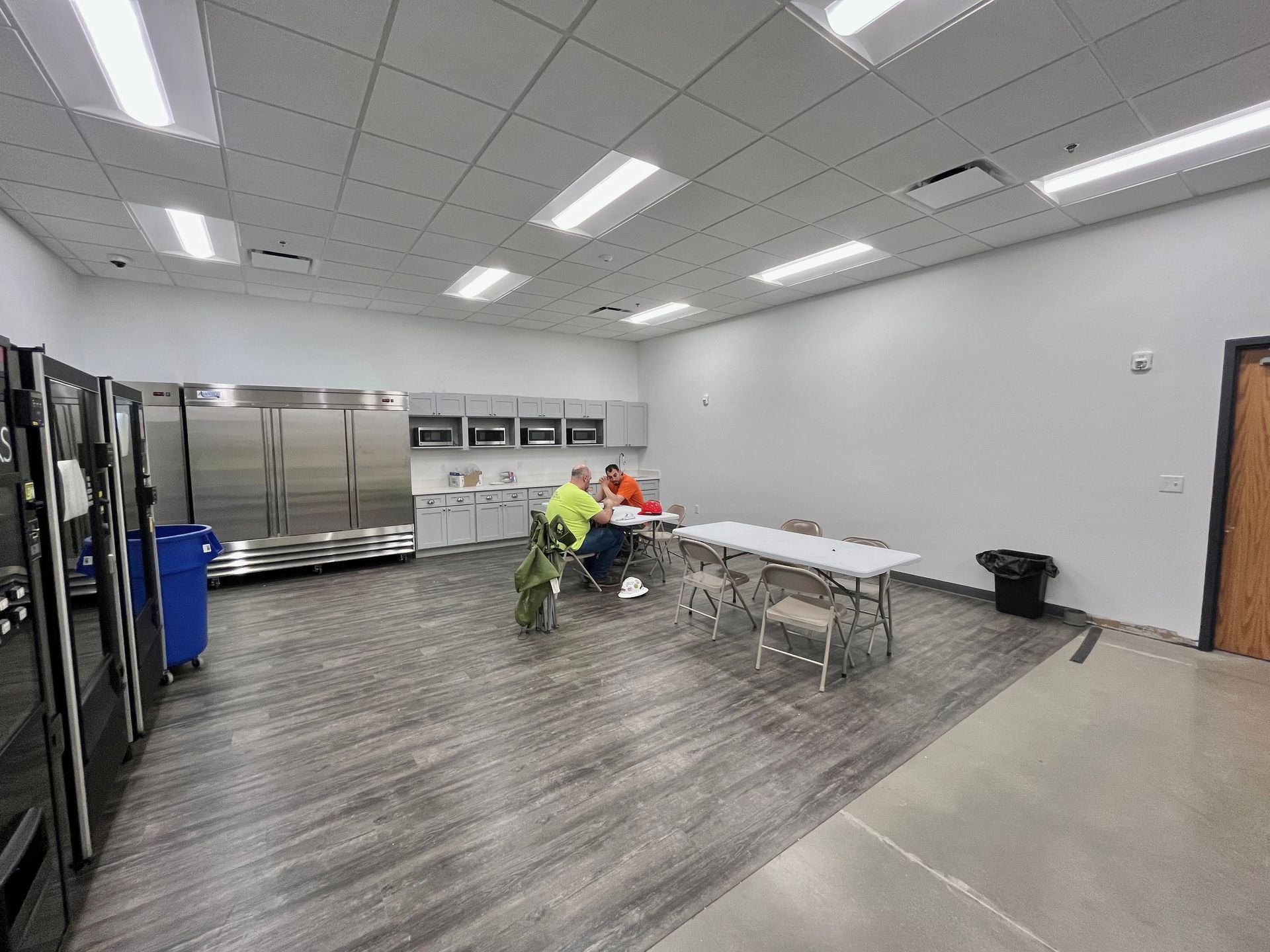 Two people sit at a table in a bright break room, near refrigerators, a sink, and a trash can.