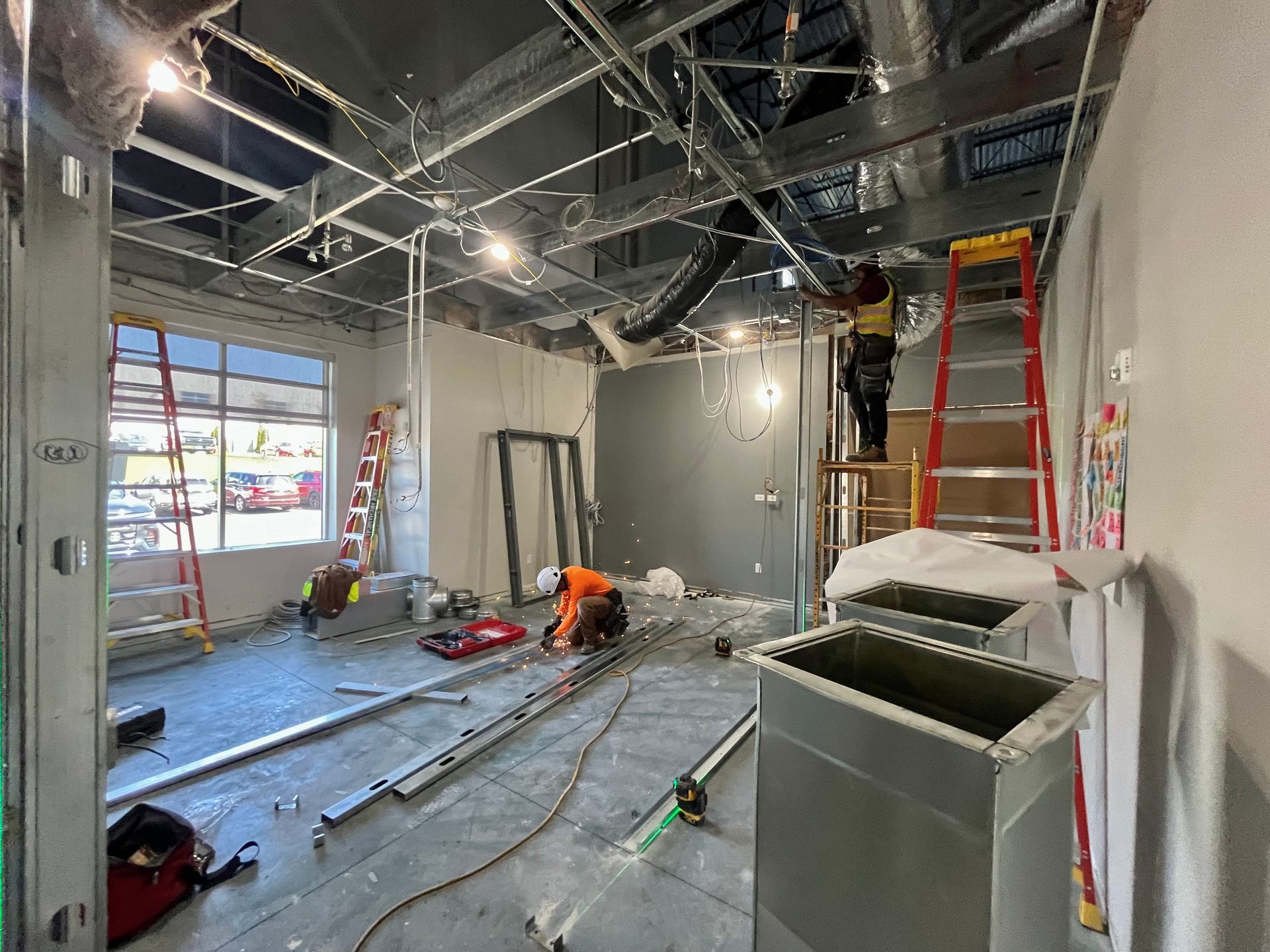 Construction site: workers installing ceiling components, ductwork, and electrical wiring. Interior shot with ladders.
