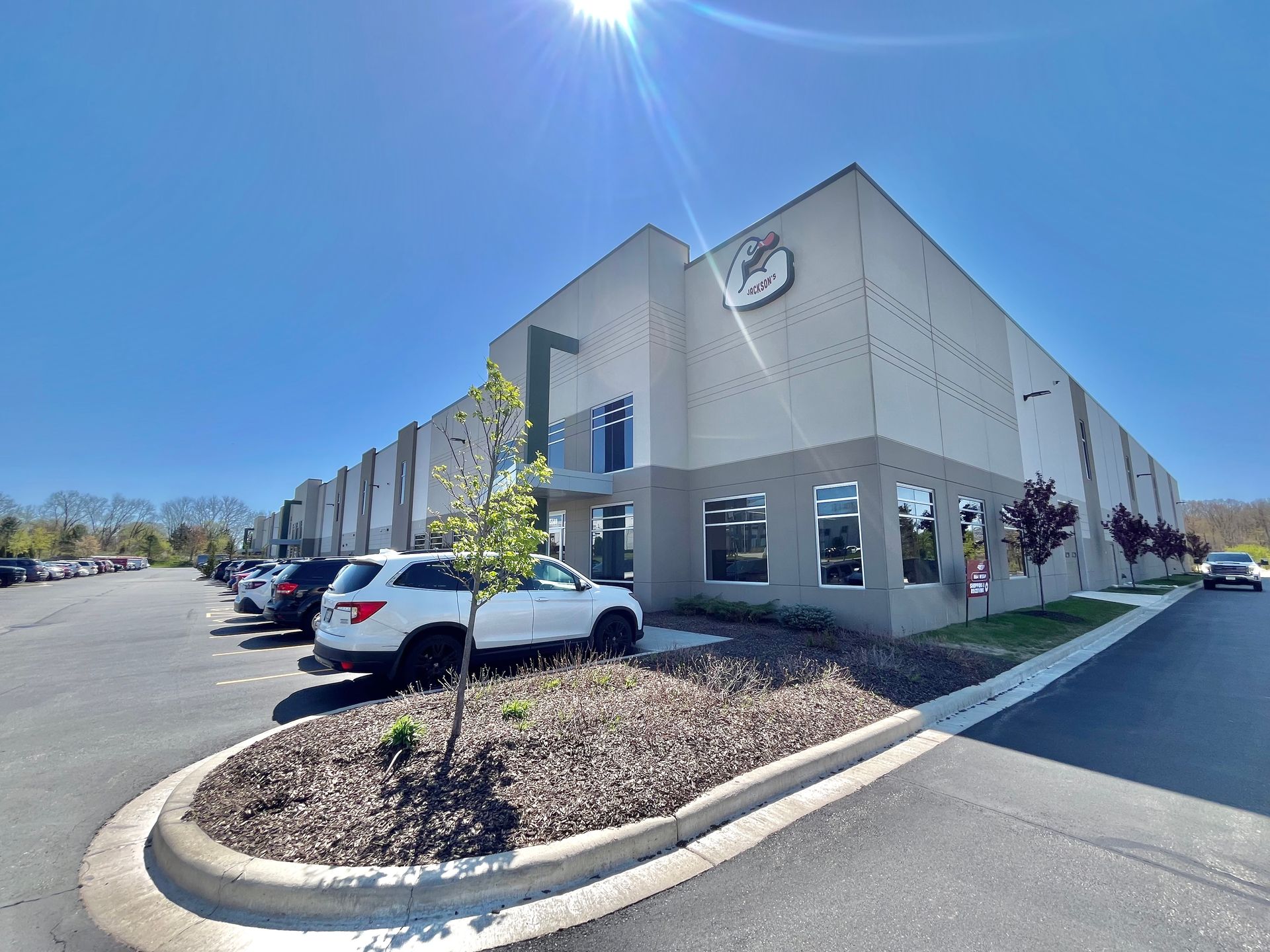 Exterior of a two-story beige industrial building with parked cars and a blue sky.