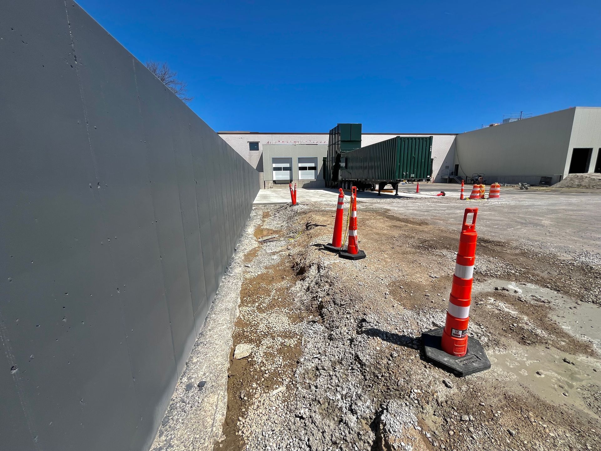 Construction zone with grey wall, orange cones, gravel, and a truck against a building under a blue sky.