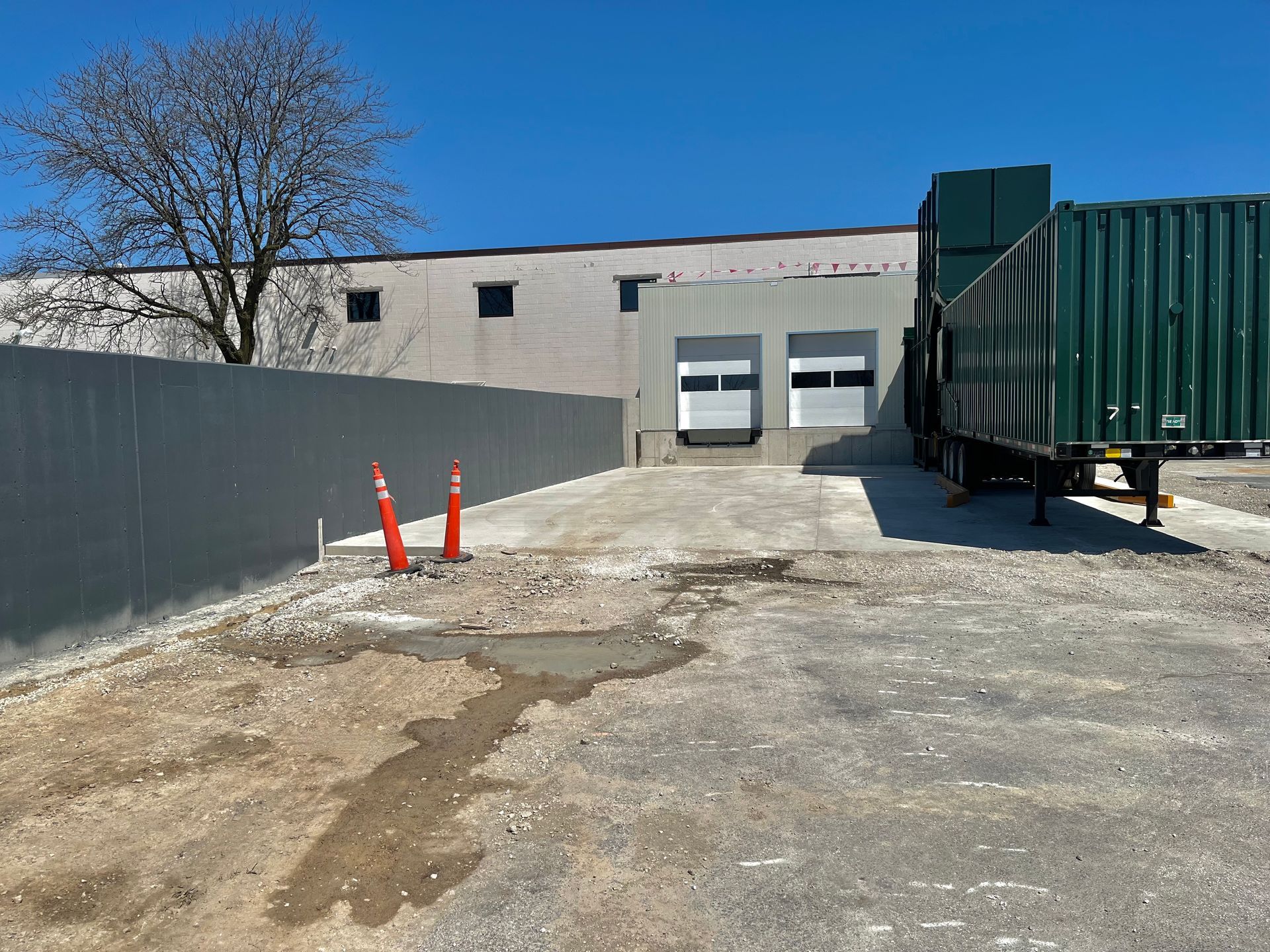 A loading dock with a parked trailer, two orange cones, and a paved ramp against a building under a blue sky.