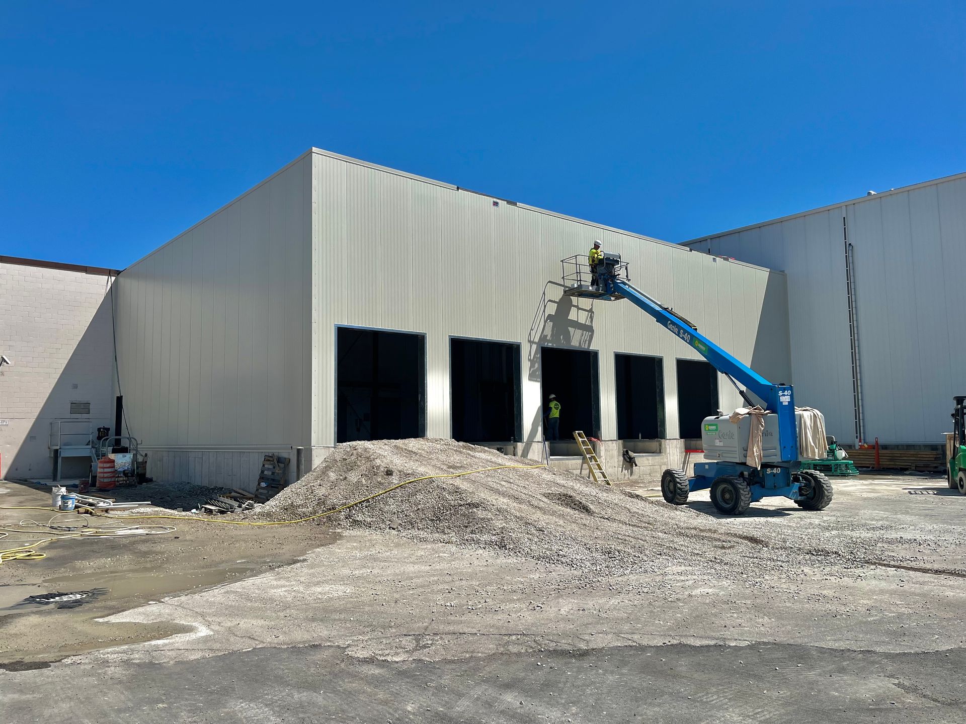 Construction site: workers on lift installing siding on a large, light-colored industrial building with loading docks.