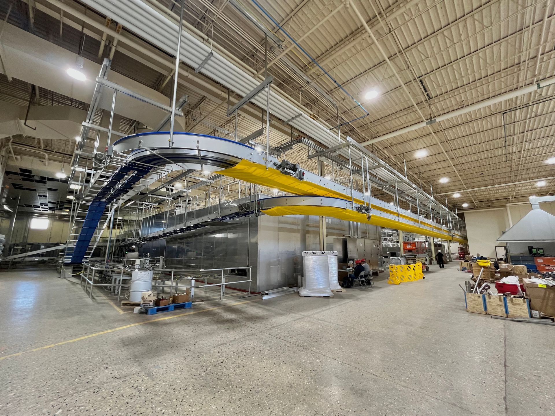 Factory interior with elevated conveyor system; large machinery with yellow and blue supports.