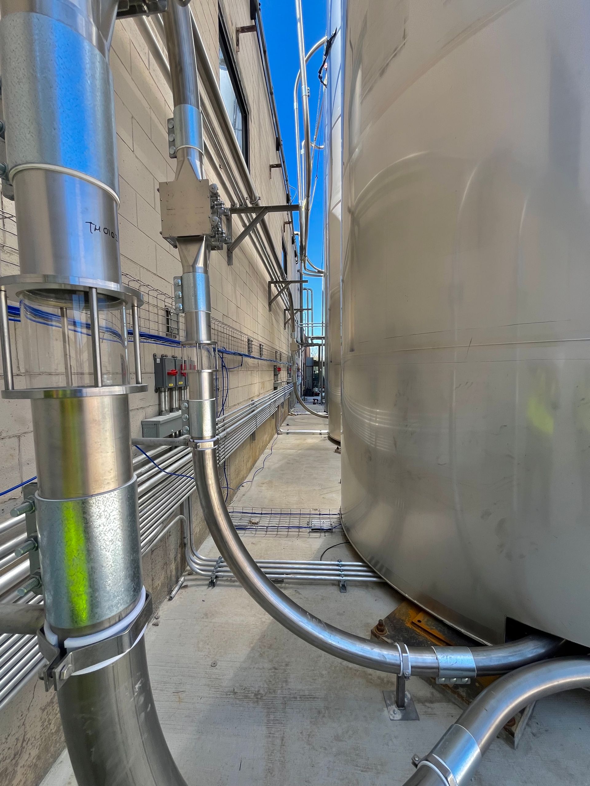 Industrial scene with metal pipes and a large tank against a wall, under a bright blue sky.