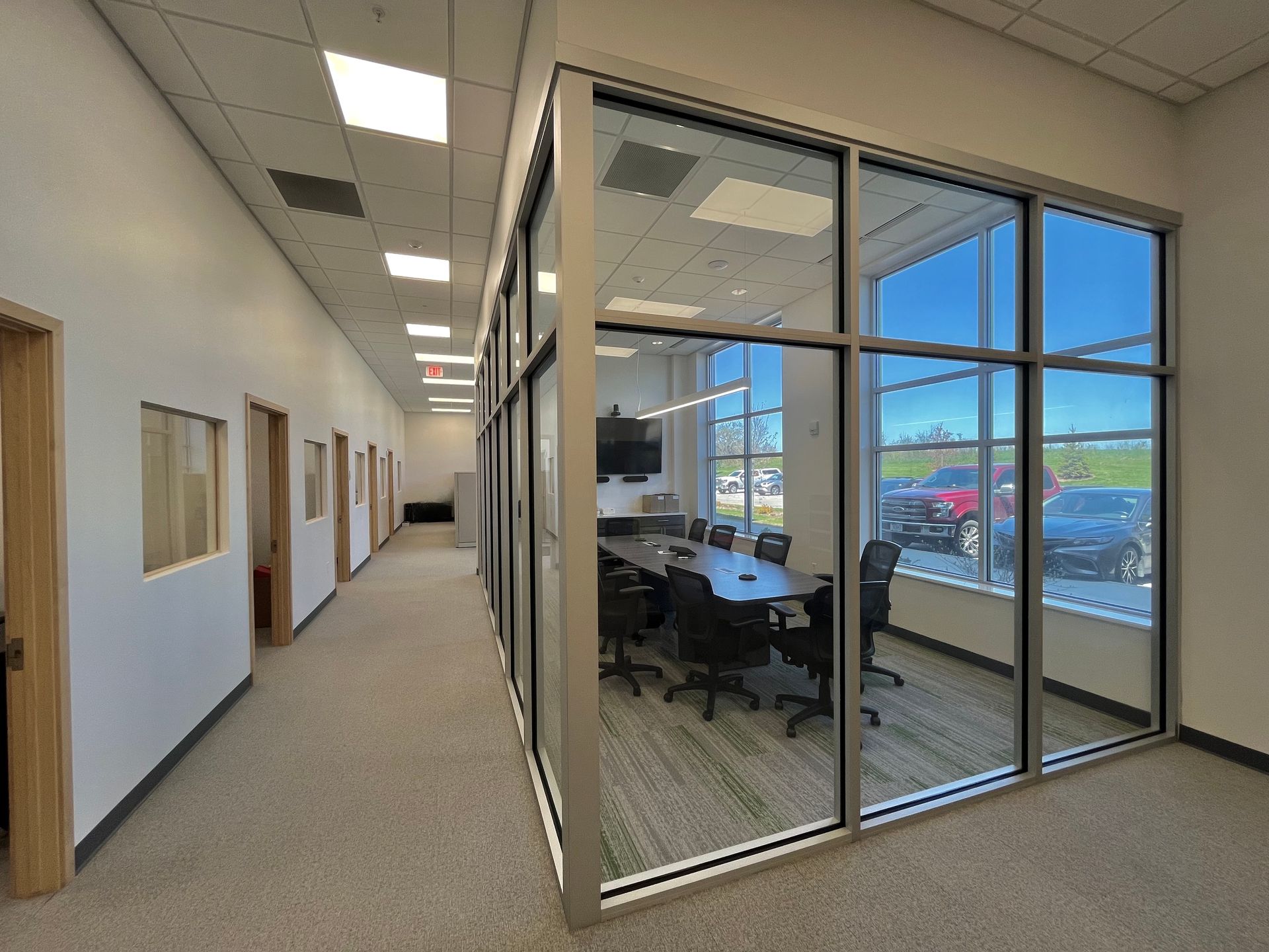 Hallway with glass-walled conference room. Interior view with a table, chairs, and windows showing an outdoor scene.