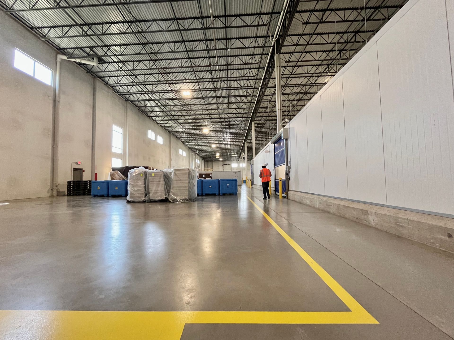 Warehouse interior with high ceiling, concrete floor, yellow line, and two people walking near white wall.