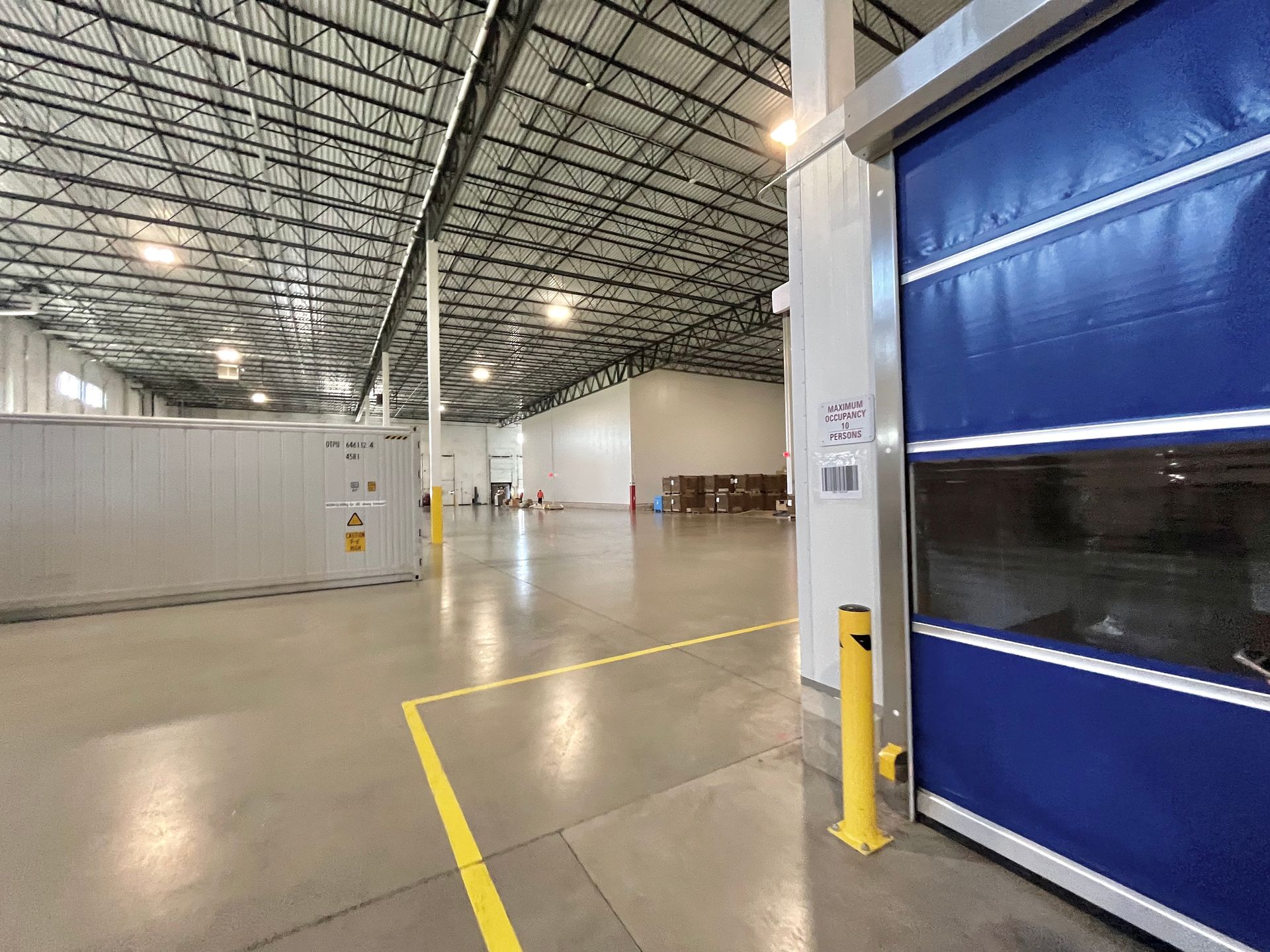 Warehouse interior with blue door, high ceiling, concrete floor, yellow lines and poles.