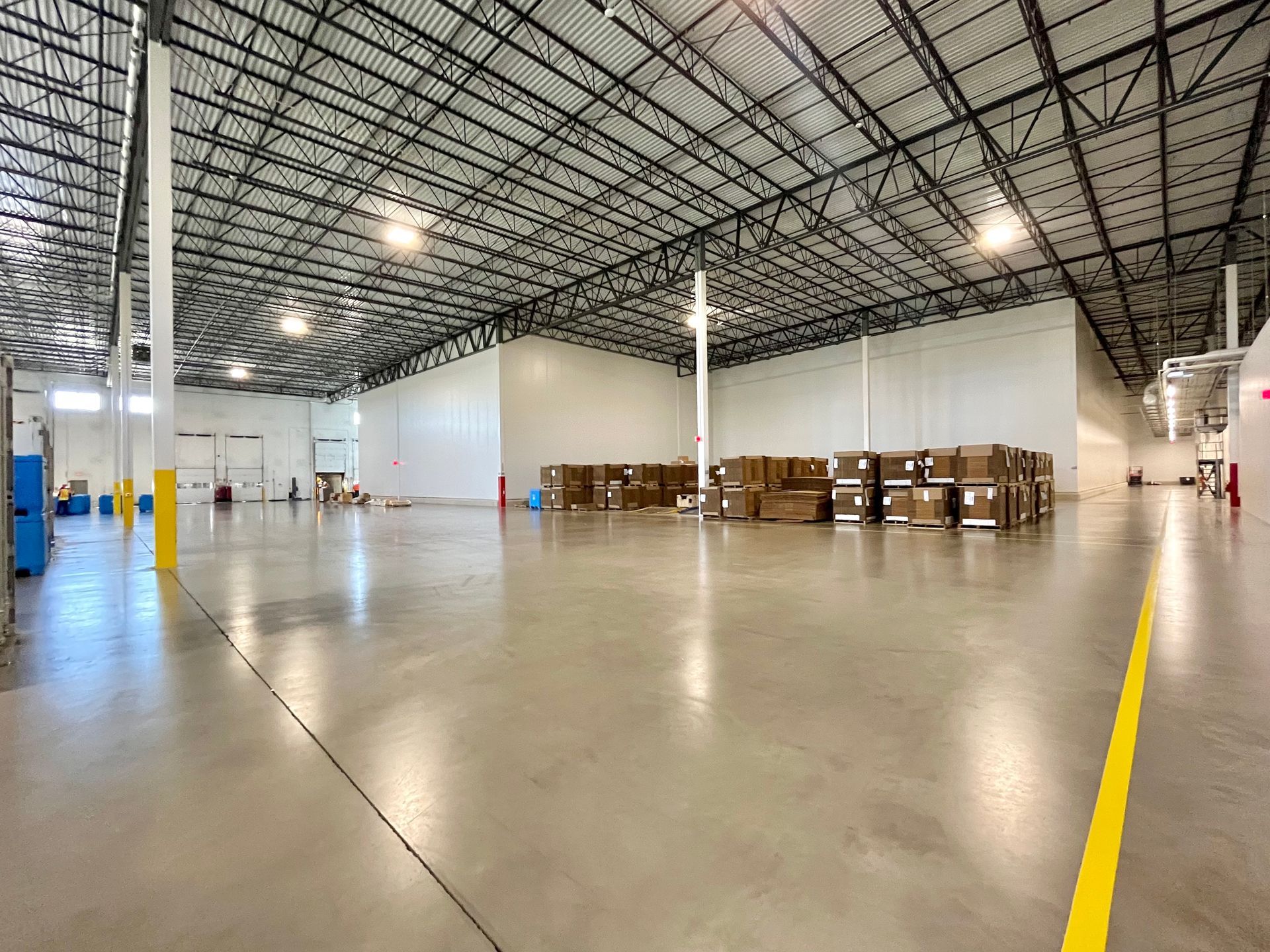 Empty warehouse interior with concrete floor, metal truss ceiling, and stacks of boxes.