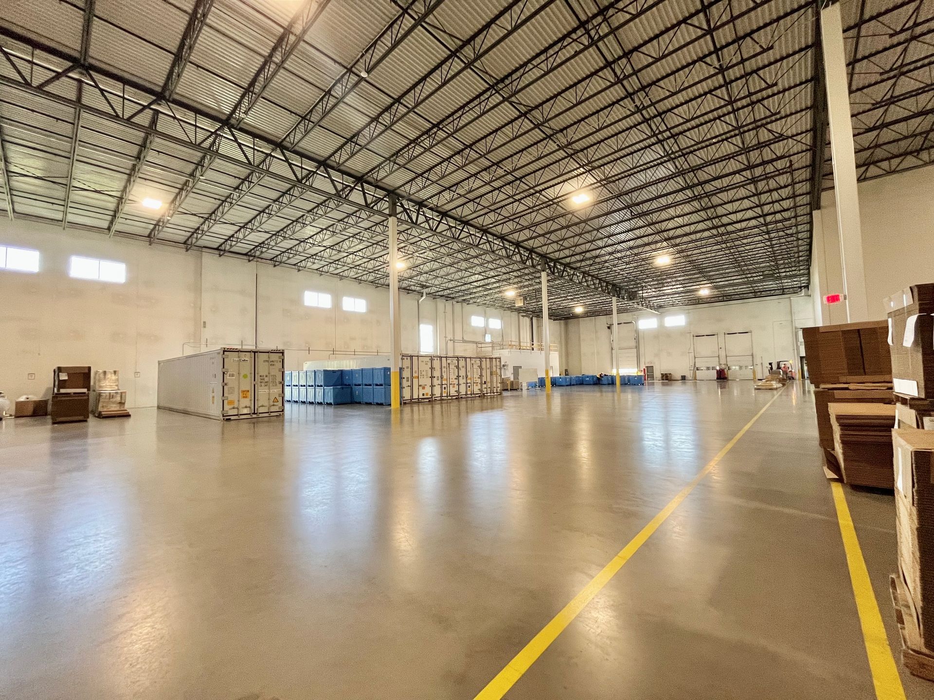 Interior of a large, empty warehouse with containers and a shiny concrete floor under a metal beam ceiling.