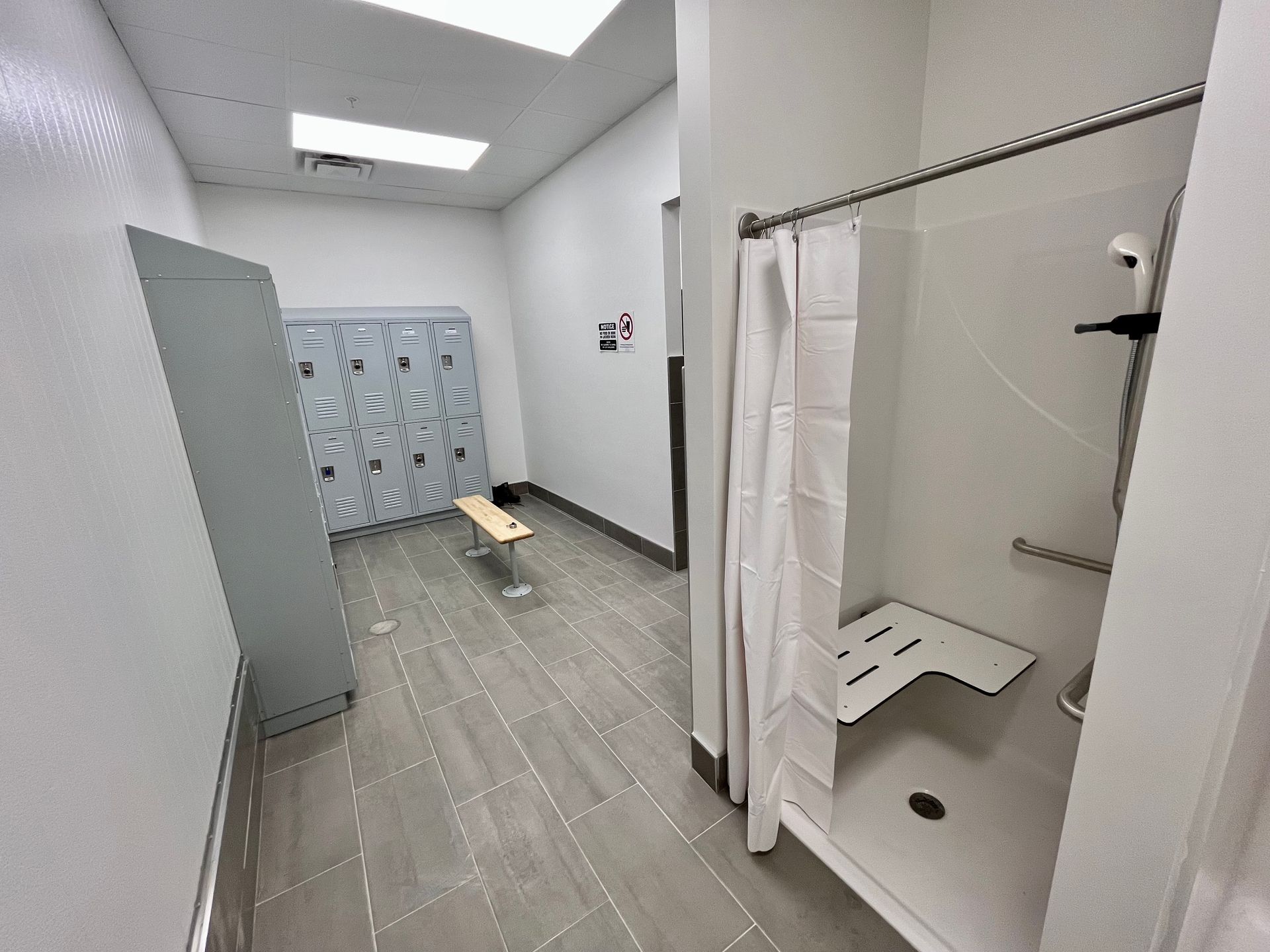 Empty locker room with shower stall. Gray lockers, bench, and floor. White shower with curtain and seat.