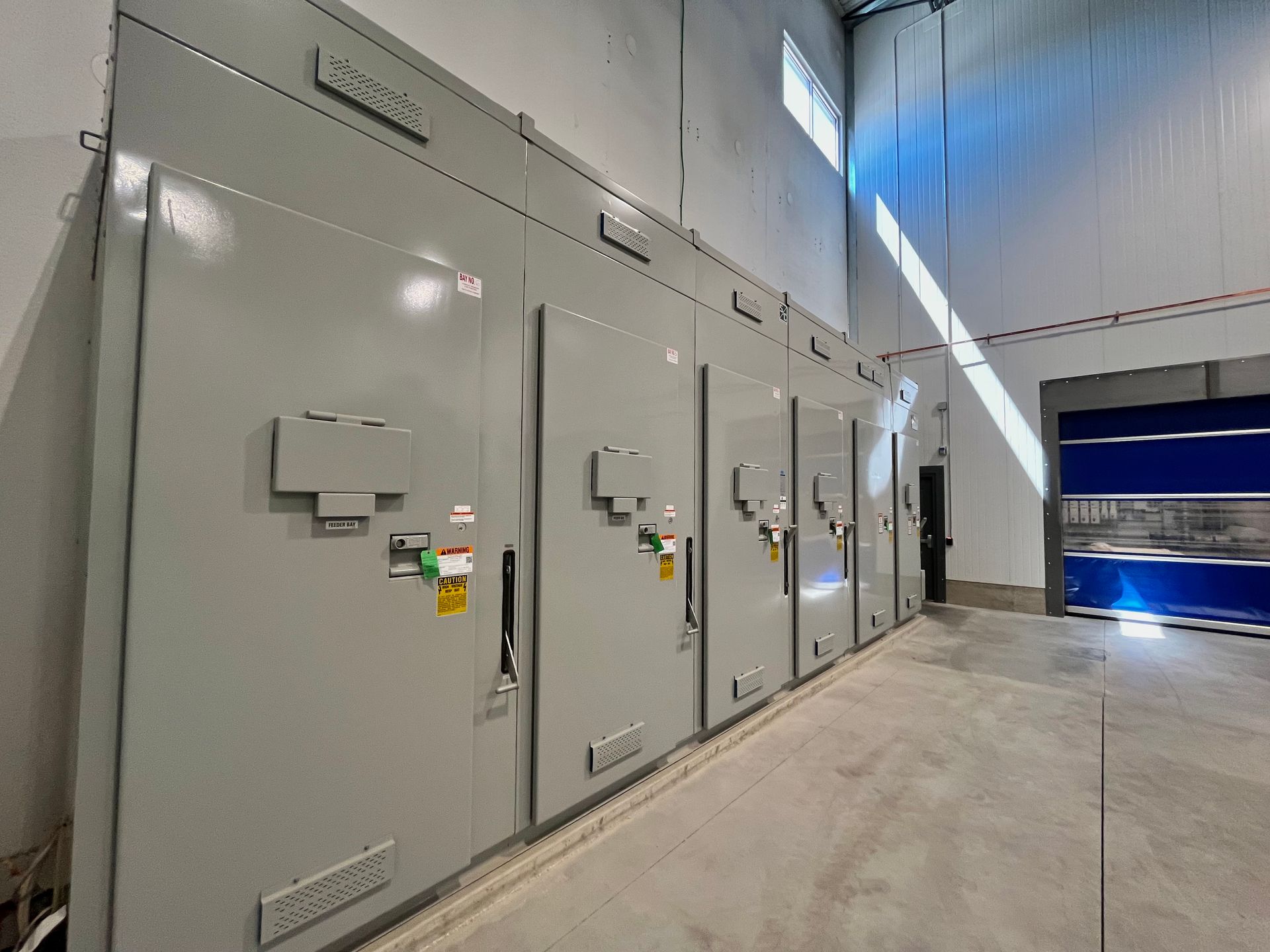 Gray electrical control panels in a bright industrial room with a concrete floor and a blue door.