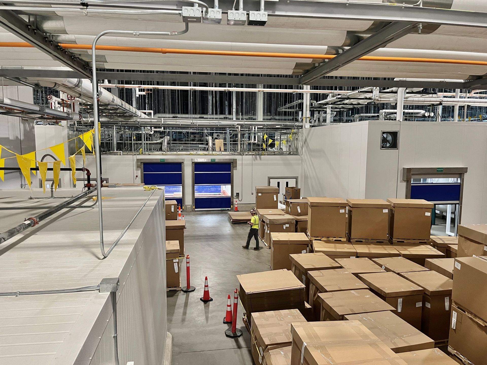 Warehouse interior with stacked cardboard boxes, two blue loading doors, and a worker in a yellow vest.