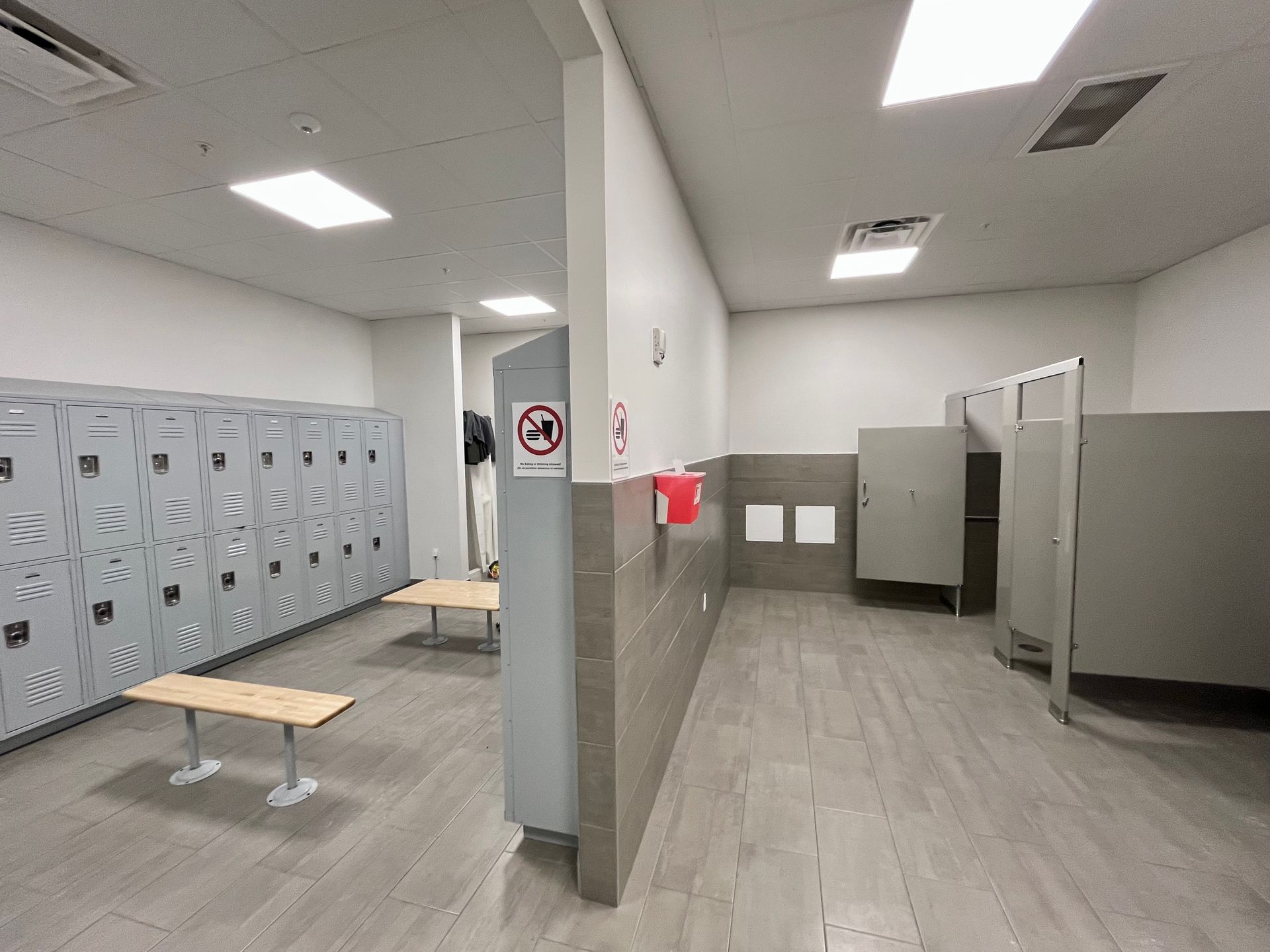 A locker room with rows of gray lockers, benches, stalls, and a no-smoking sign.