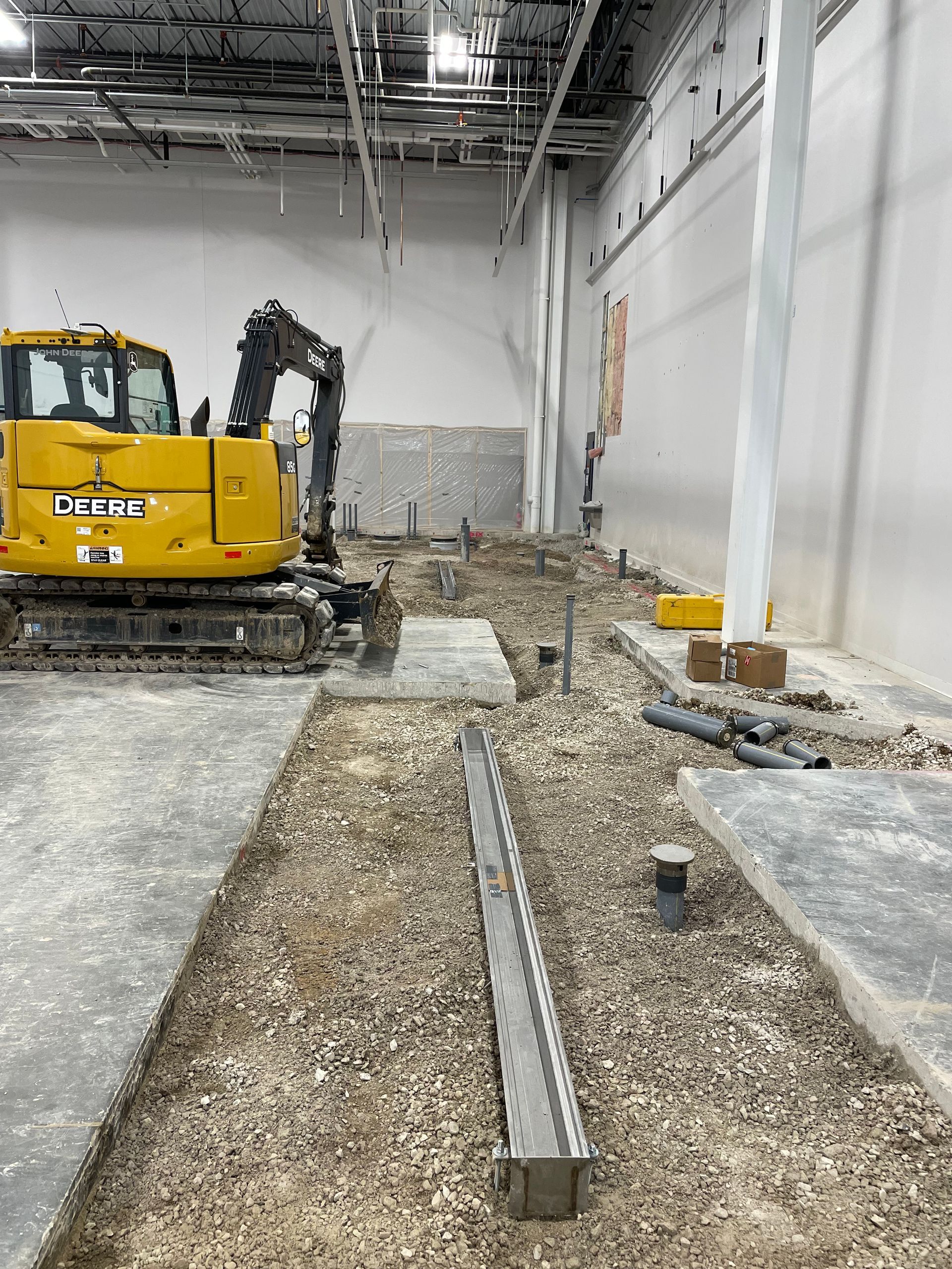 Construction site: Excavator in large building, trench with drain, exposed plumbing.