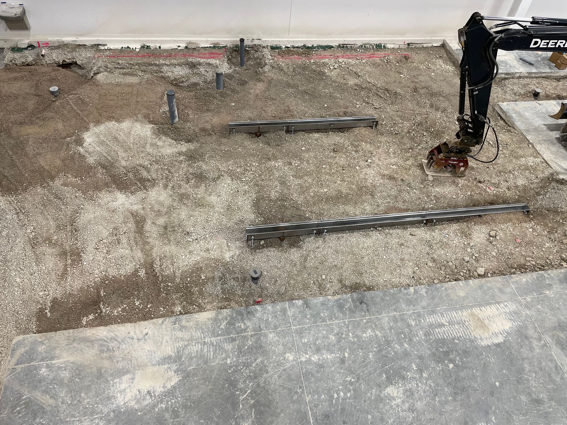 Construction site. Concrete floor with exposed piping and gravel. A compactor tool stands nearby.
