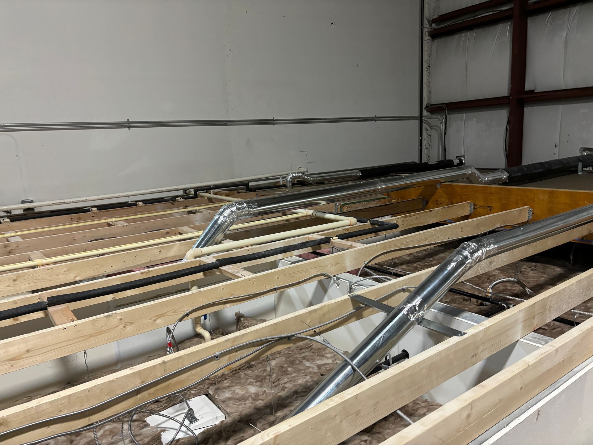 Interior view of a building under construction. Wooden beams and metal ducts on the floor. Wires and insulation are visible.
